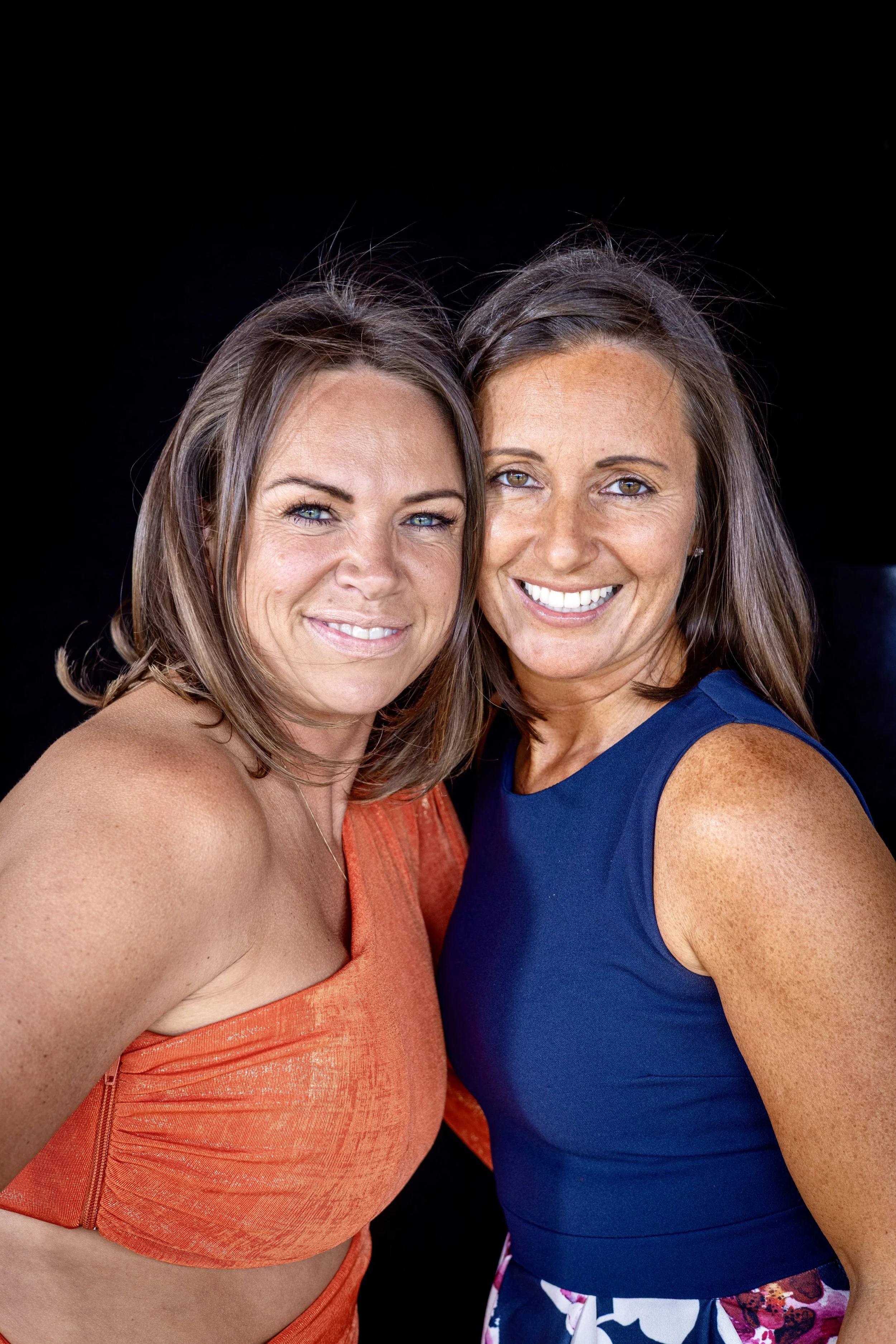 Two women standing close together against a black background, smiling at the camera.