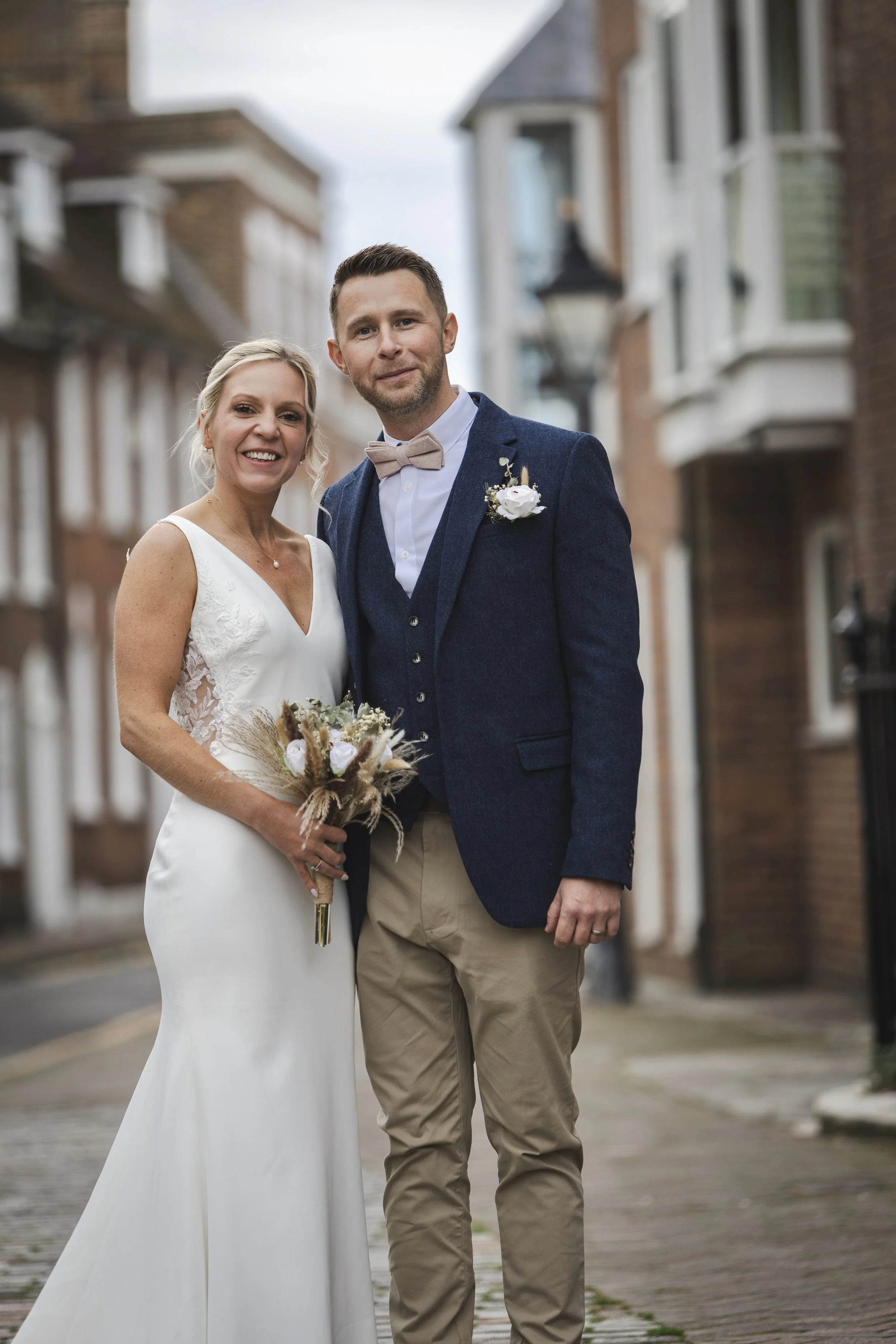 A bride and groom standing together on a street, smiling for a photo during their wedding. The bride is wearing a white wedding dress and holding a bouquet, while the groom is dressed in a navy blazer, beige pants, and a beige bow tie.
