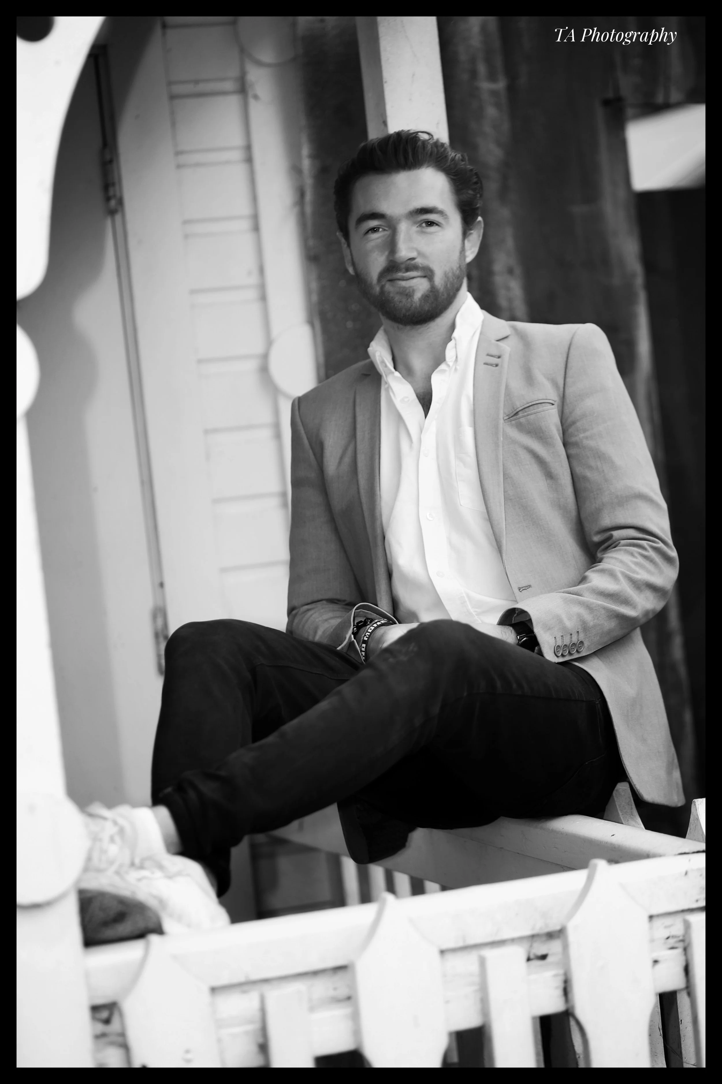 A black and white photo of a man with a beard and styled hair, dressed in a blazer and white shirt, sitting with crossed legs in an outdoor urban setting. There is a brand name "TA Photography" in the top right corner.
