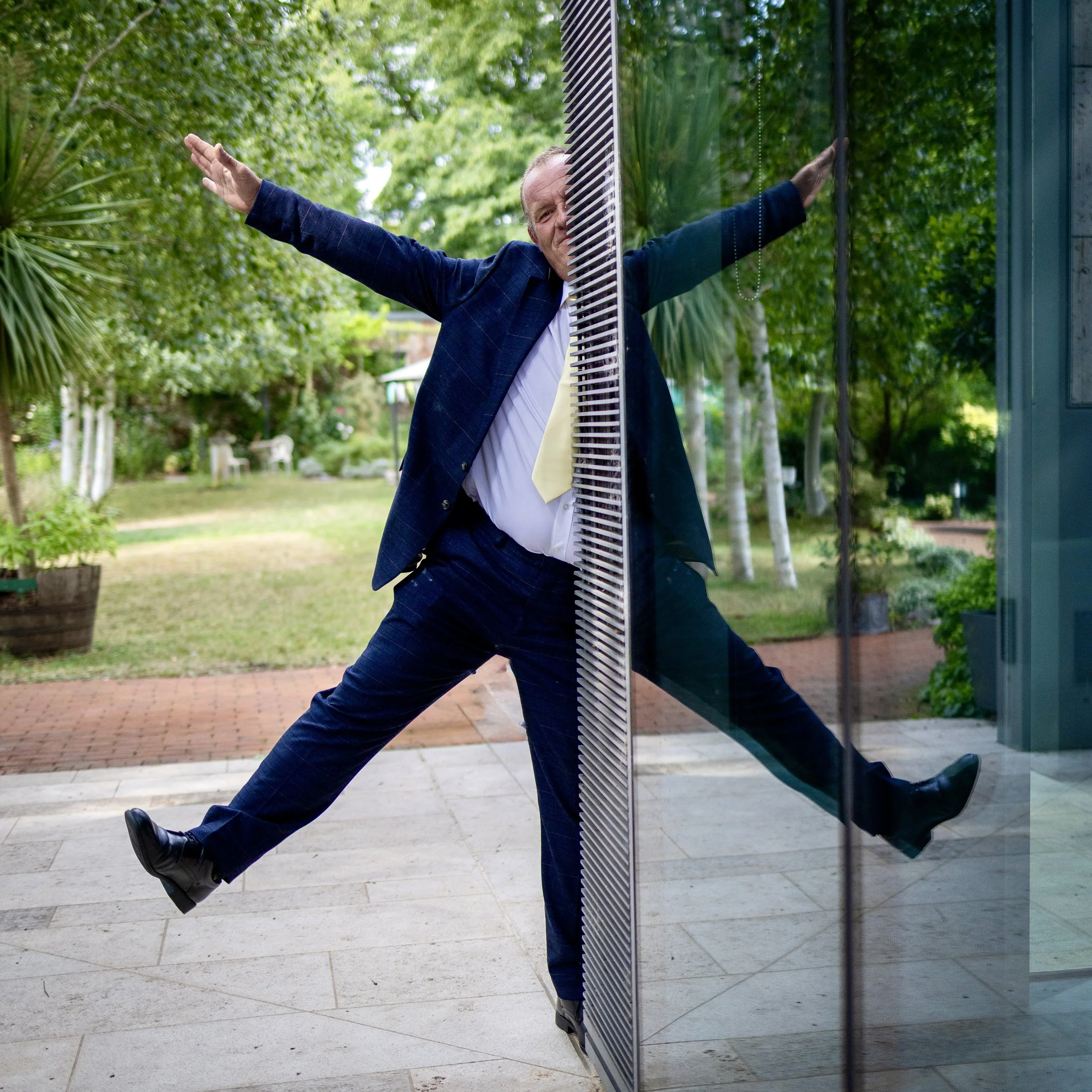 A man in a business suit standing in front of a glass door with a smile, arms outstretched, one leg raised, and the other on the ground, outdoors with trees and plants in the background.