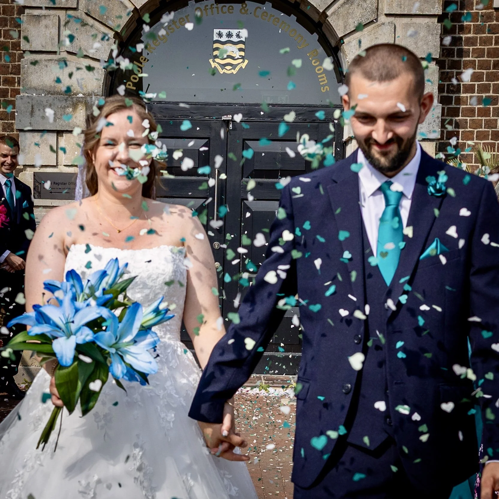 A newly married couple holding hands and smiling, surrounded by falling confetti, outside a building with a sign that reads 'Poole Register Office & Ceremony Rooms,' during their wedding celebration.