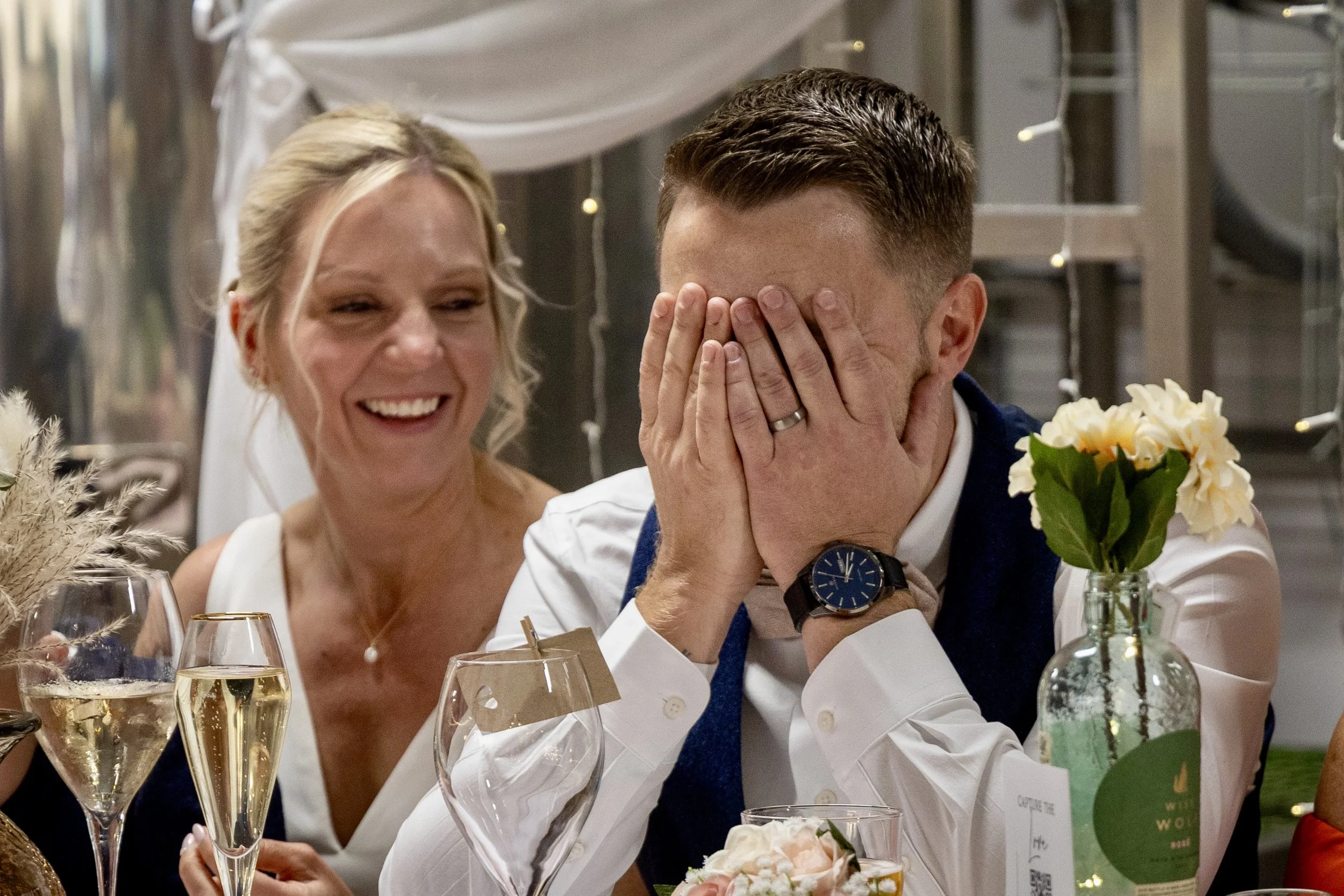 A woman with blond hair and a man with short brown hair are at a celebration, with the woman smiling and the man covering his face with his hands. There are glasses of champagne, flowers, and decorations on the table.
