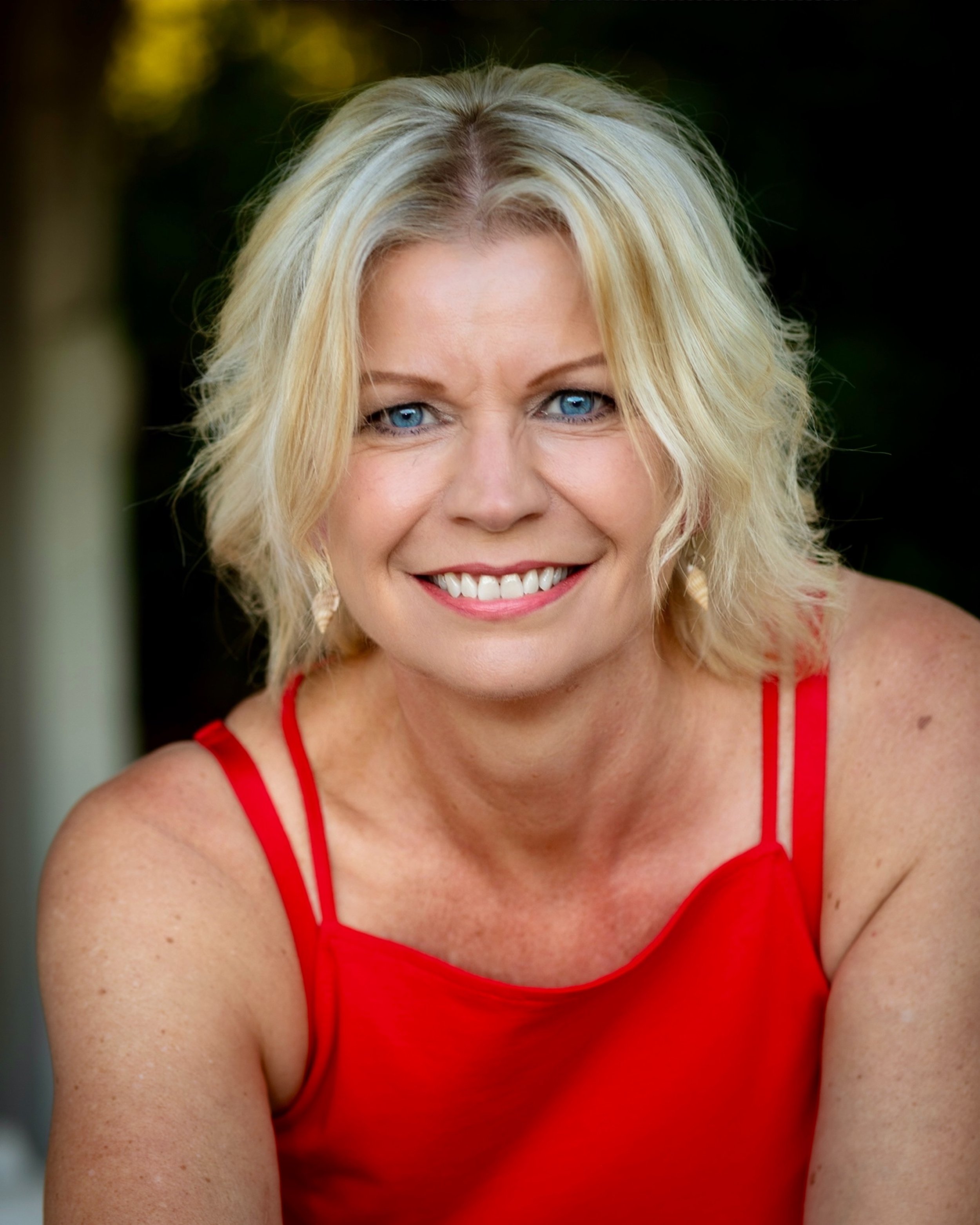Head and shoulders of a middle-aged woman with blonde wavy hair, blue eyes, and a bright smile, wearing a red sleeveless top, set outdoors with a blurred natural background.