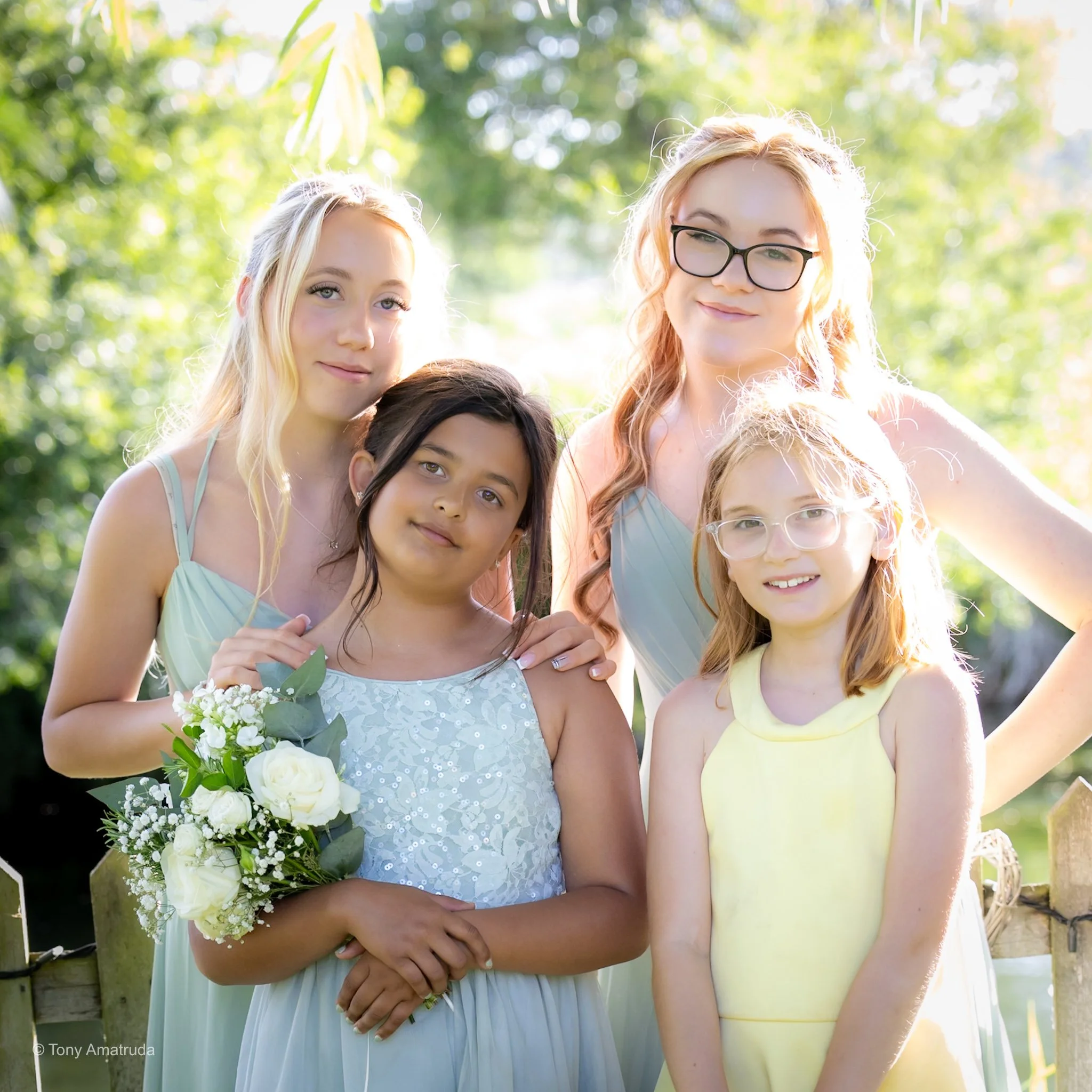 A group of women, including a bride holding a bouquet of white flowers, standing outdoors in a sunny, green environment.