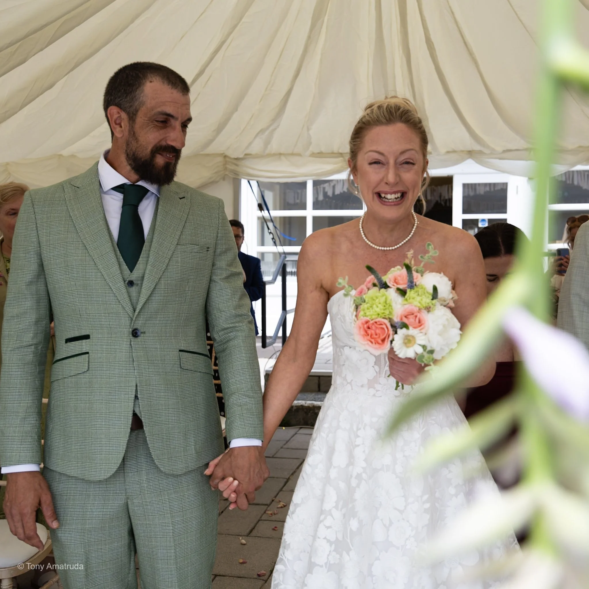 A bride and groom holding hands during their wedding inside a tent, with the bride holding a bouquet of pink and white flowers, and both dressed in formal wedding attire.