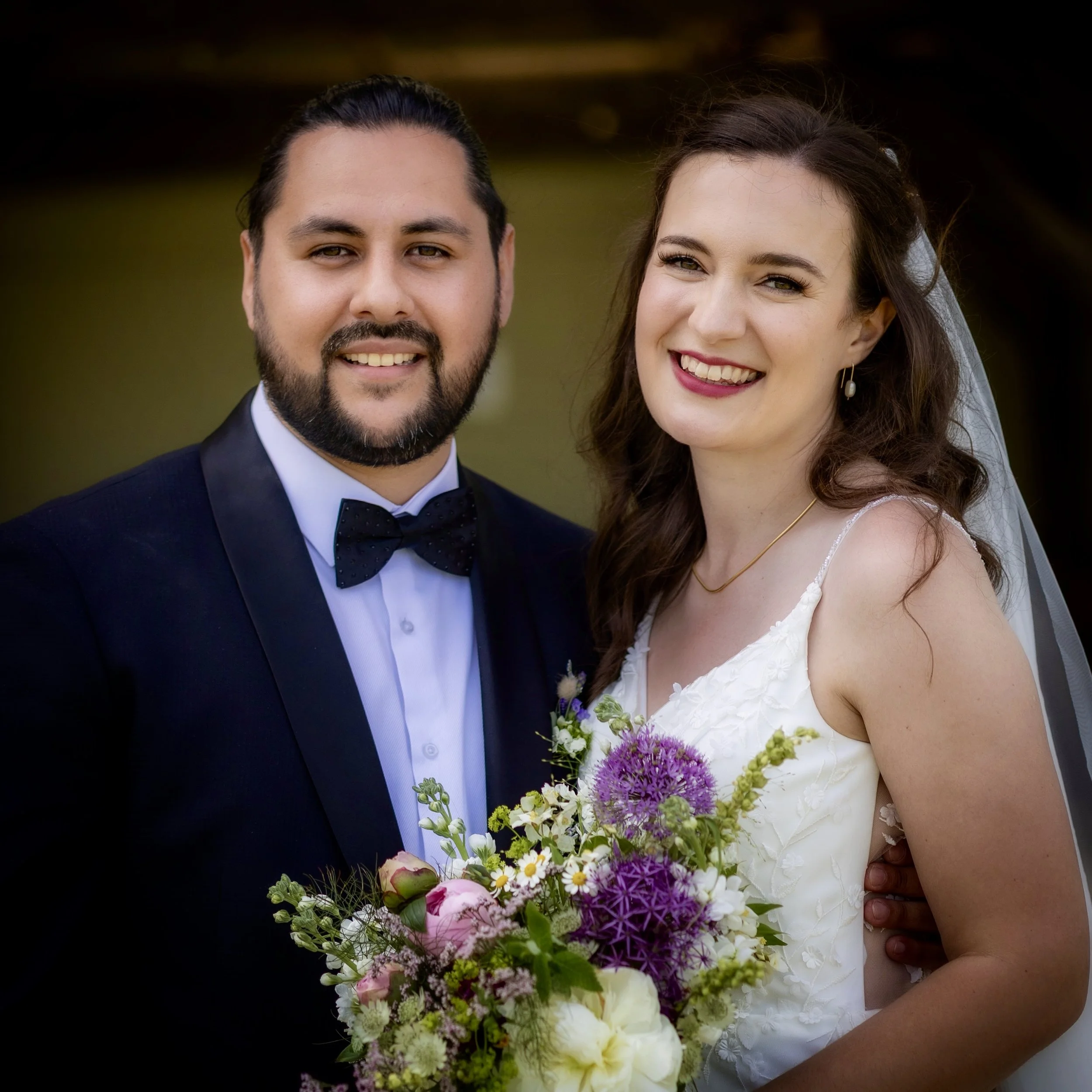 A bride and groom on their wedding day, smiling outdoors, with the bride holding a colorful bouquet of flowers.