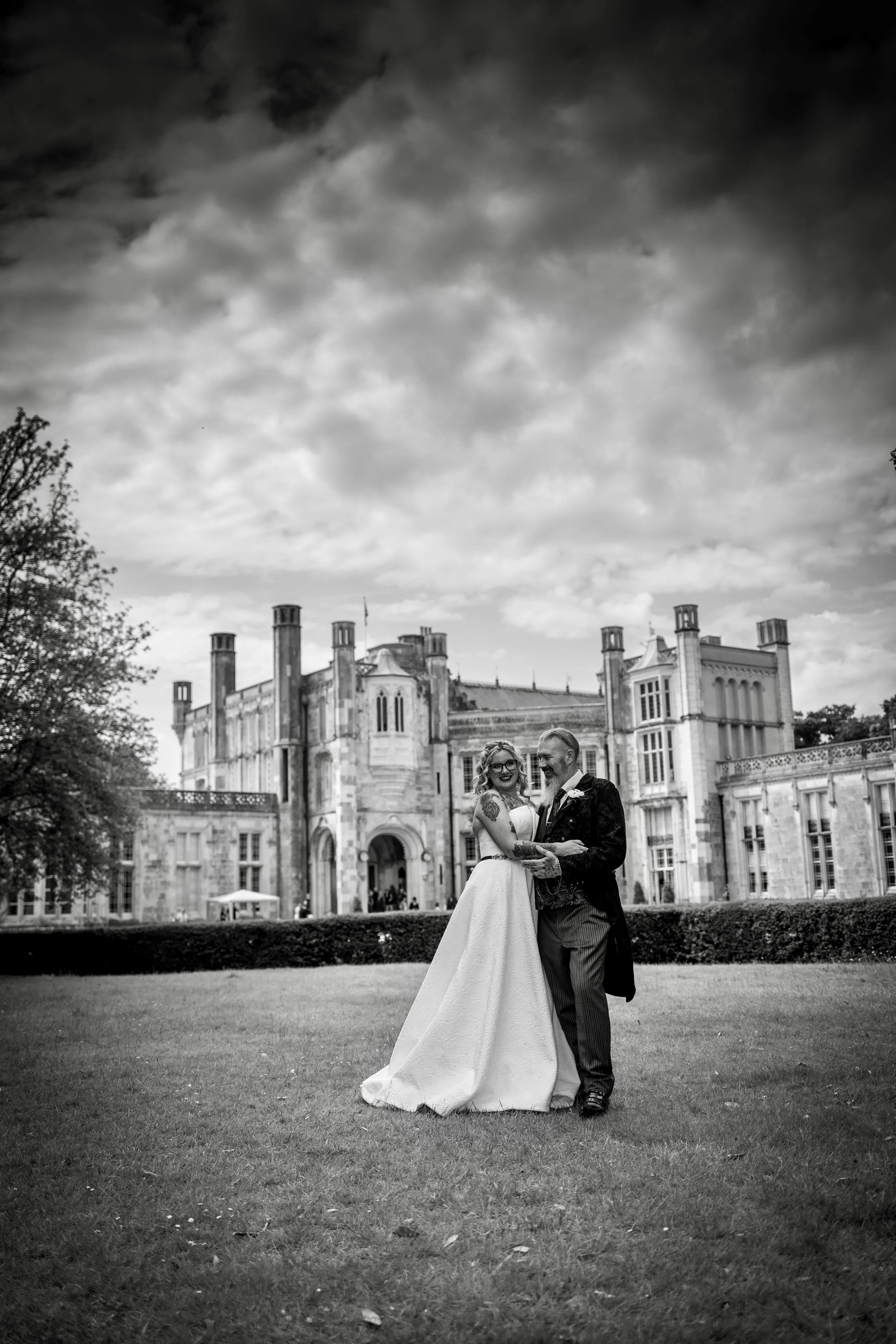 A black-and-white photo of a bride and groom dancing on the lawn outside a large, historic castle-like building with cloudy sky in the background.
