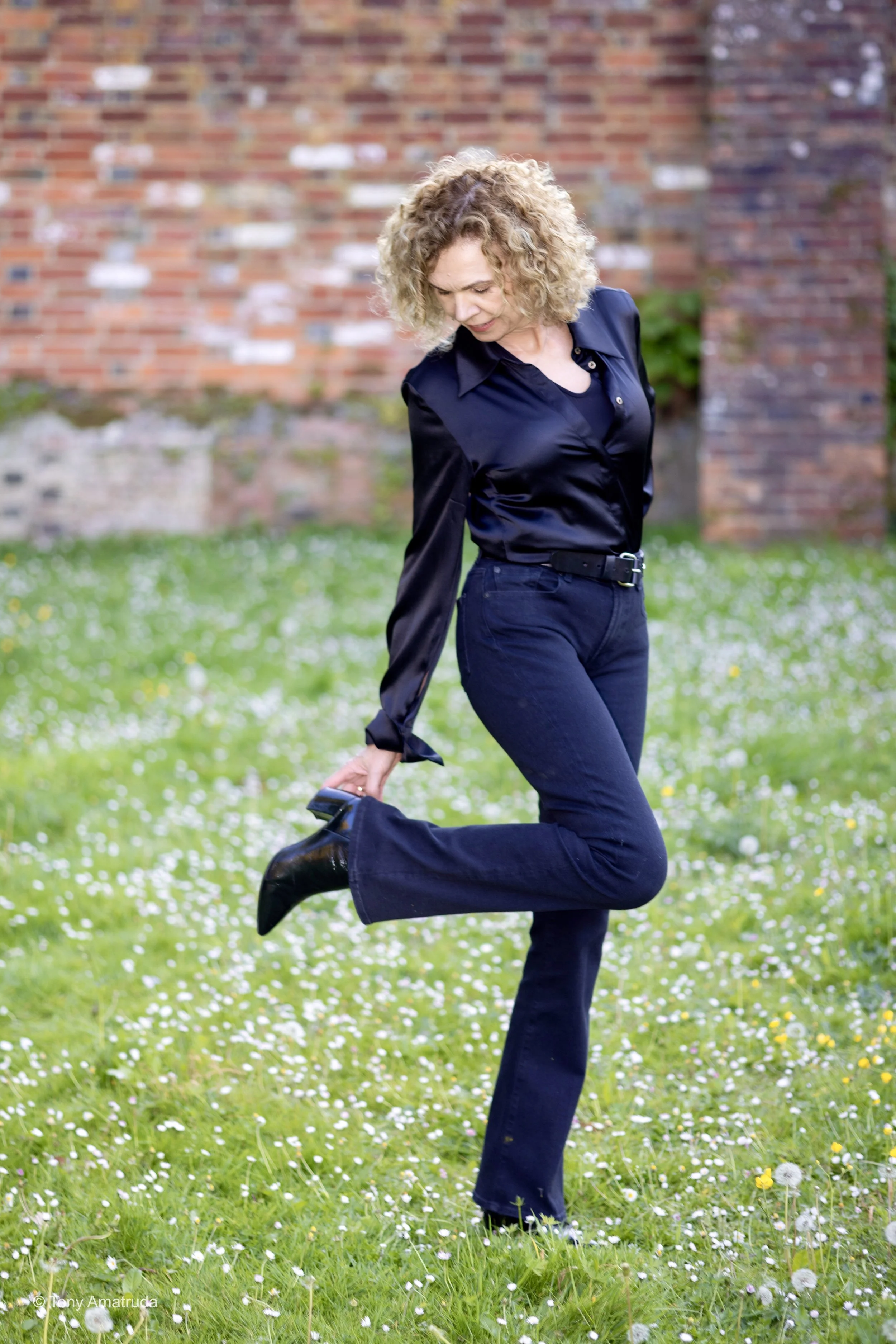A woman with curly blonde hair wearing a black shirt and black pants, standing outdoors on a grassy area with small flowers, holding one foot behind her as she balances on the other leg.