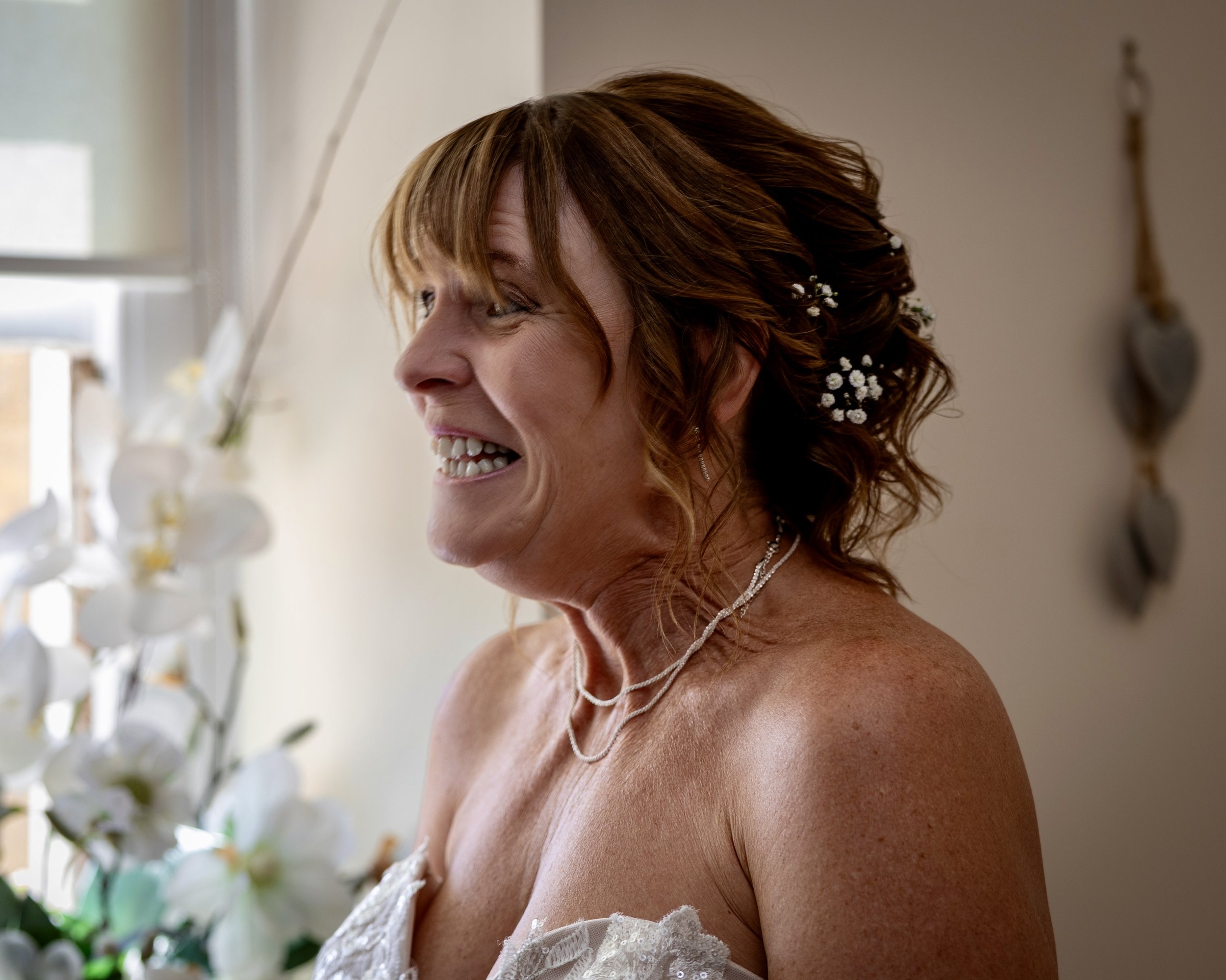 Close-up of a woman with red hair smiling, wearing a white dress with lace details and floral accessories in her hair, in a softly lit indoor setting.