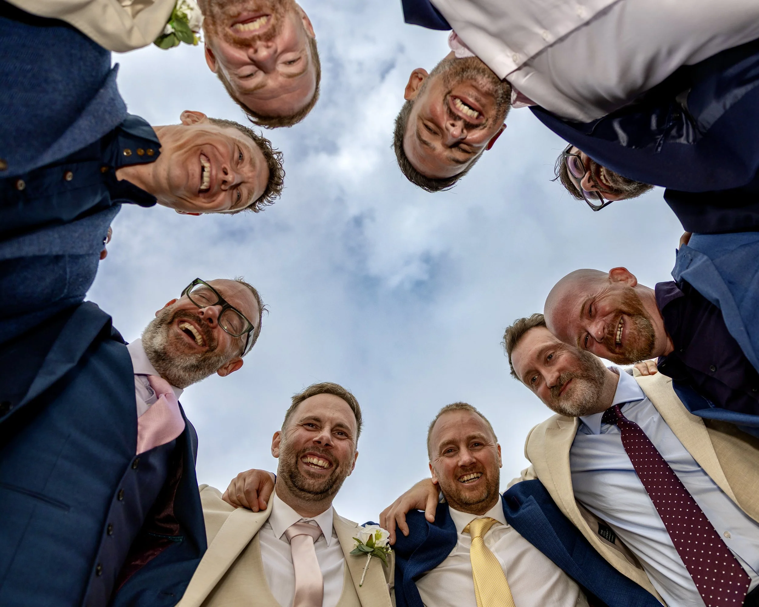 A group of men, dressed in suits and ties, standing in a circle outdoors, smiling and leaning in towards each other with their heads together, under a cloudy sky.