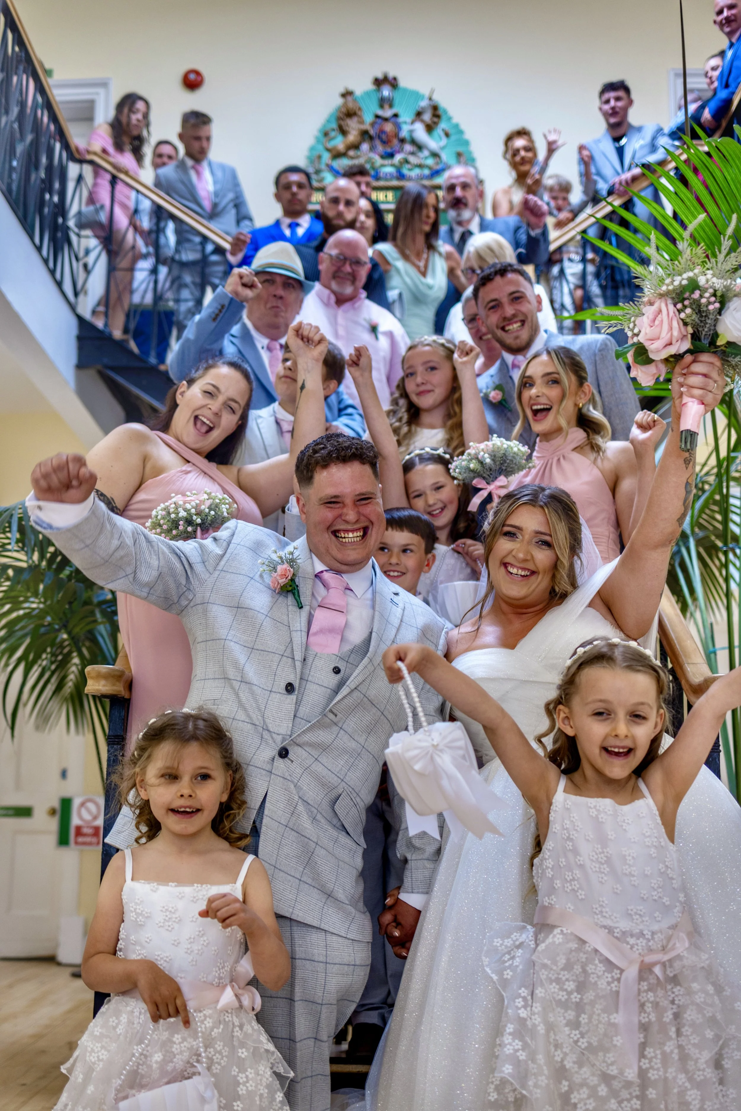 A large group of people celebrating at a wedding, with the bride and groom at the front, smiling and raising their fists. The bride is holding a bouquet, and children are in front with joyful expressions. The background features a decorative crest an