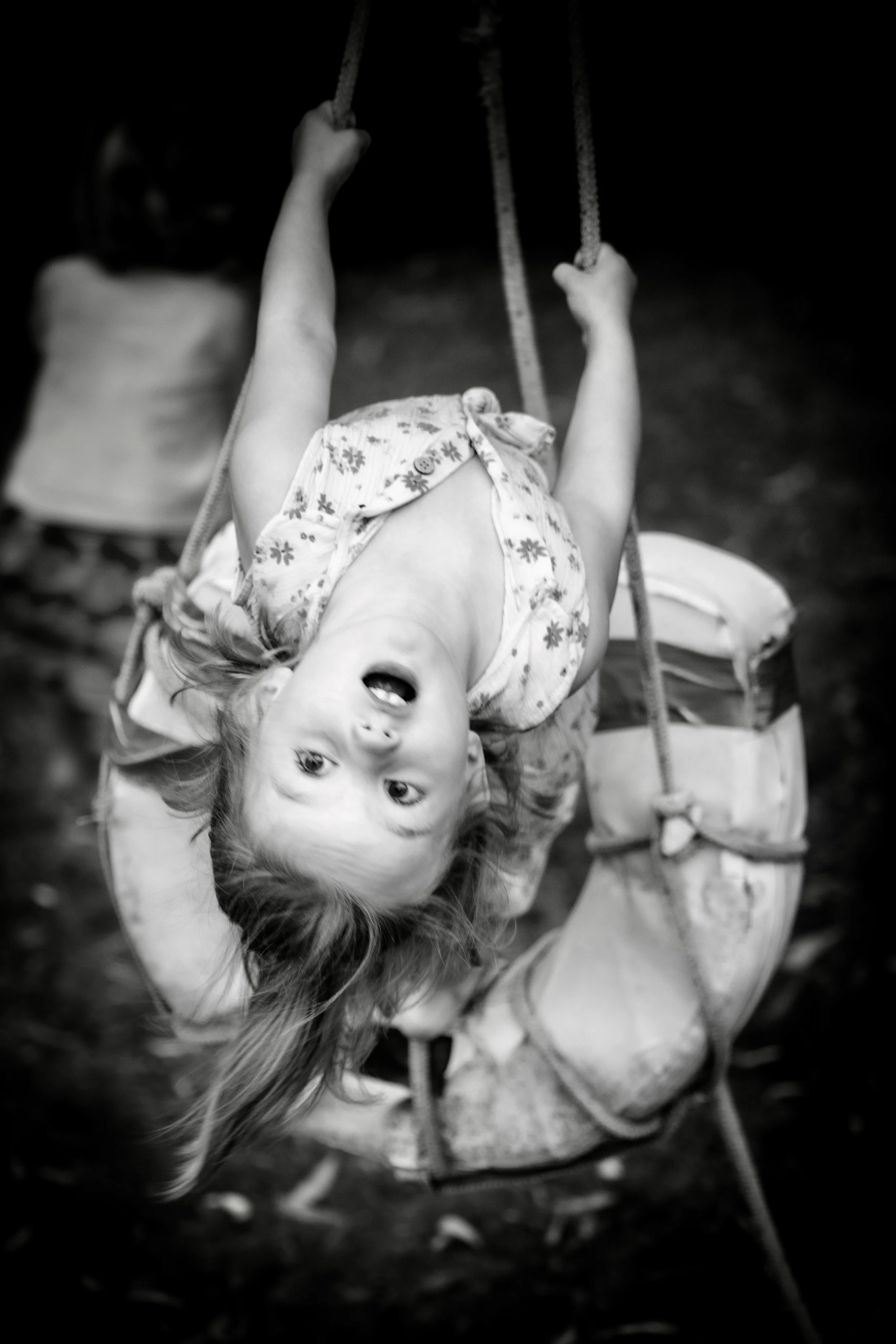 A young girl with long hair swinging upside down on a playground swing, smiling at the camera. Another child is partially visible in the background.
