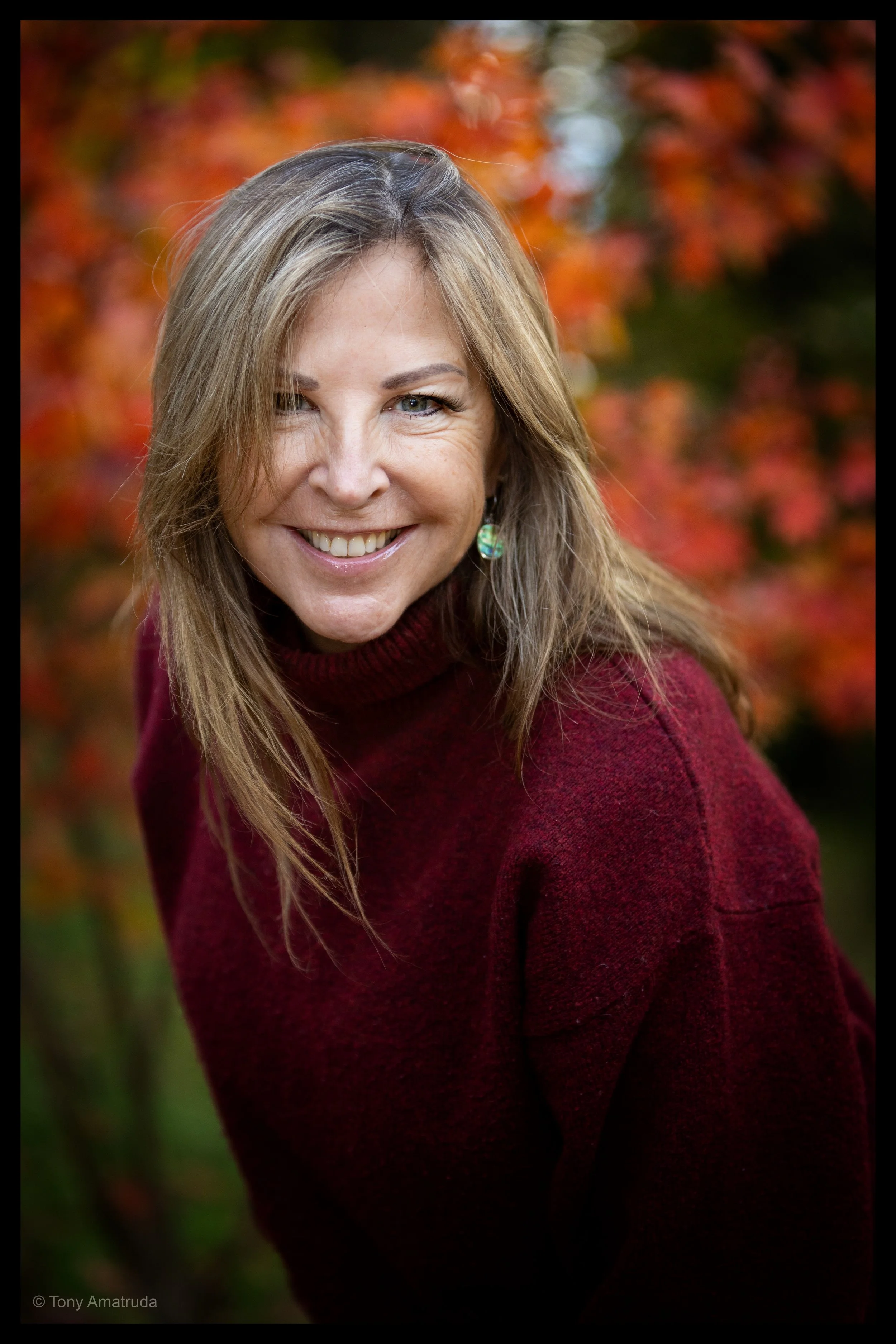 A smiling woman with light brown hair and blue eyes, wearing a maroon sweater and a green earring, outdoors with a background of fall foliage in red and orange.