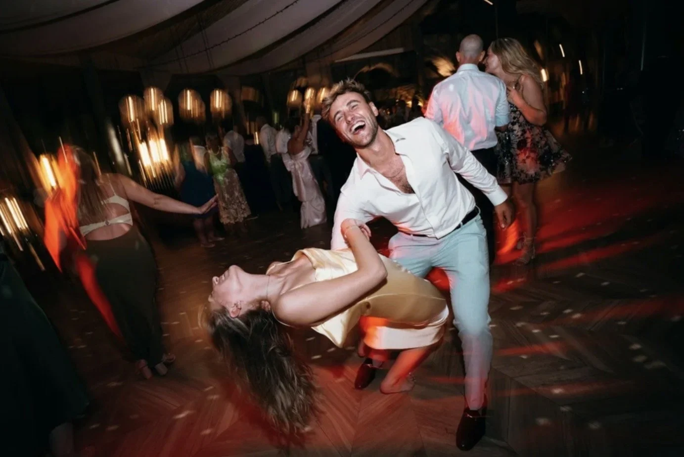 A man and woman dancing and laughing at a lively party or nightclub, with other people dancing in the background under warm lighting.