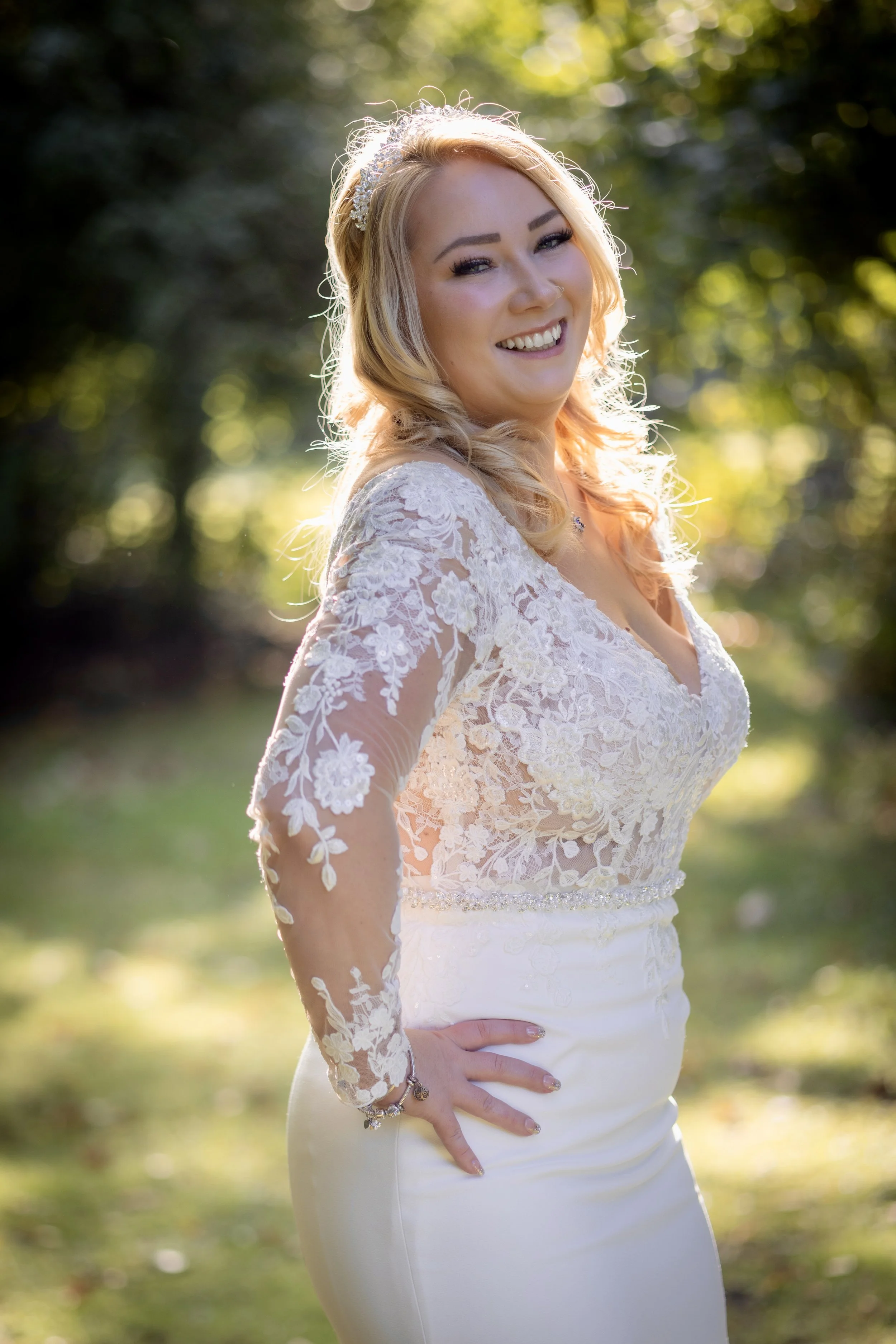 A smiling woman in a white lace wedding dress standing outdoors in a sunny, green area.