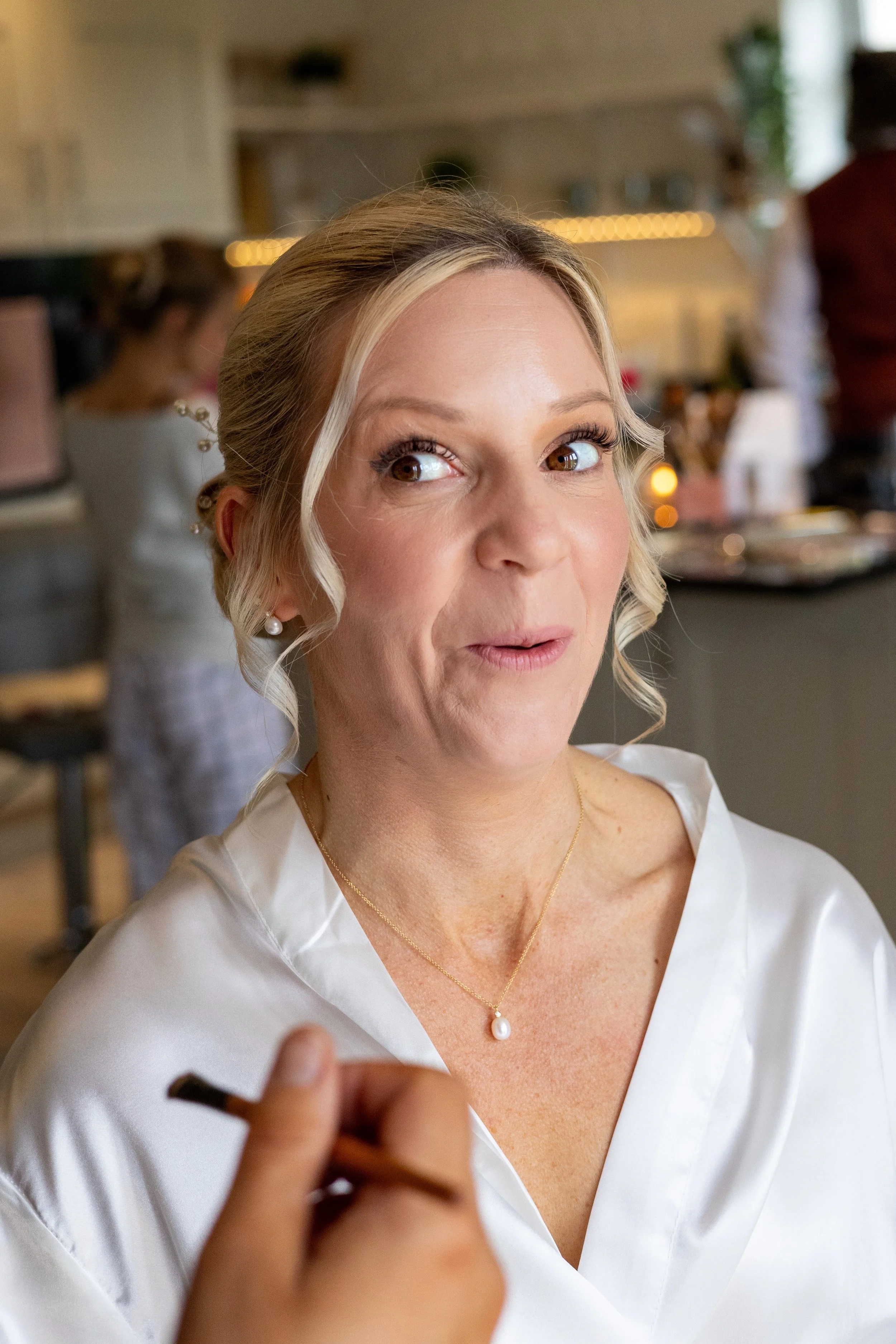 A woman with blonde hair styled in loose curls, wearing a white satin dress, pearl earrings, and a pearl necklace, appears to be in a social setting, possibly a wedding or special event, as she glances to the side while someone is taking her photogra