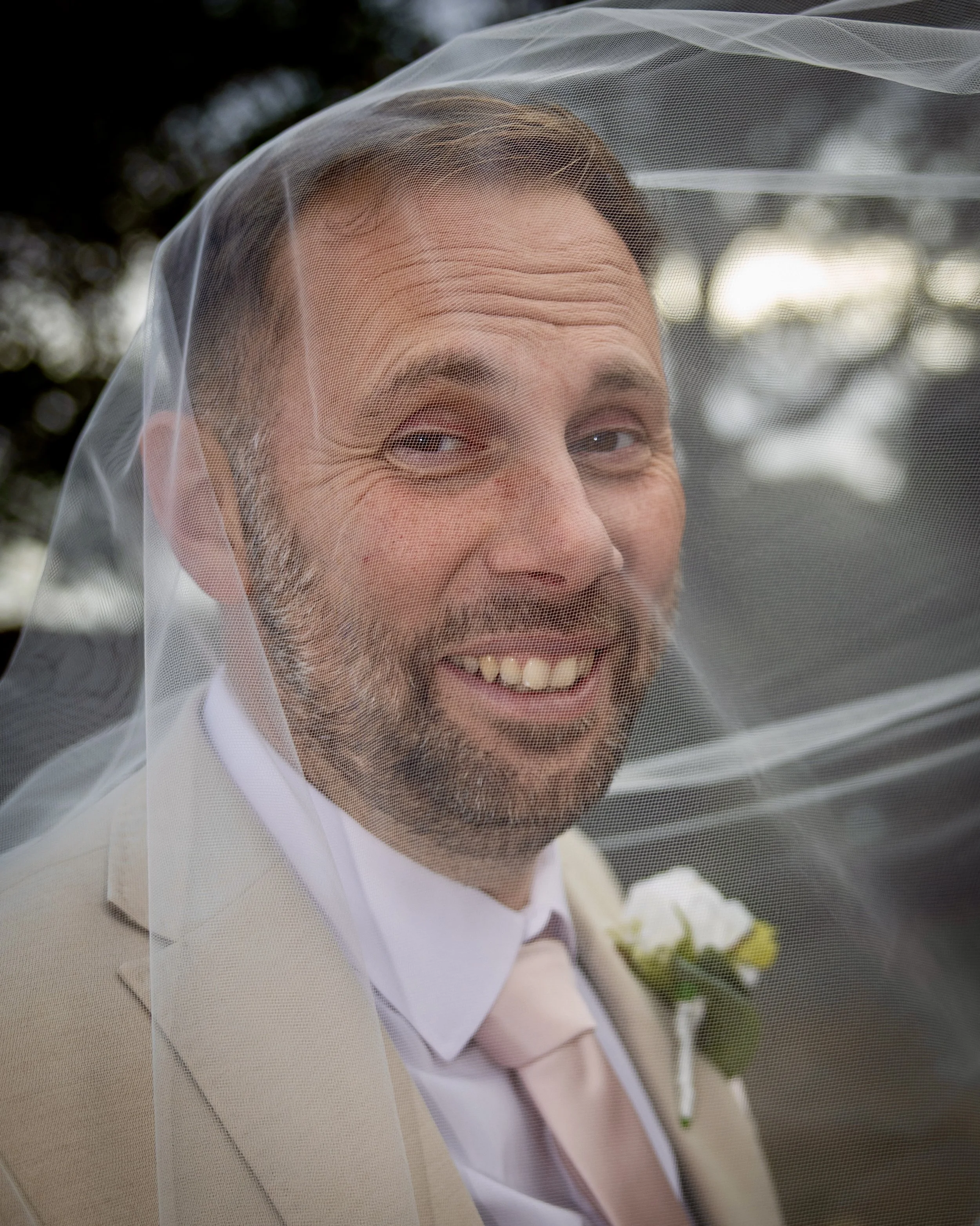 A man smiling, wearing a white tuxedo with thin black pinstripes, and a veil over his face, outdoors with blurred trees in the background.