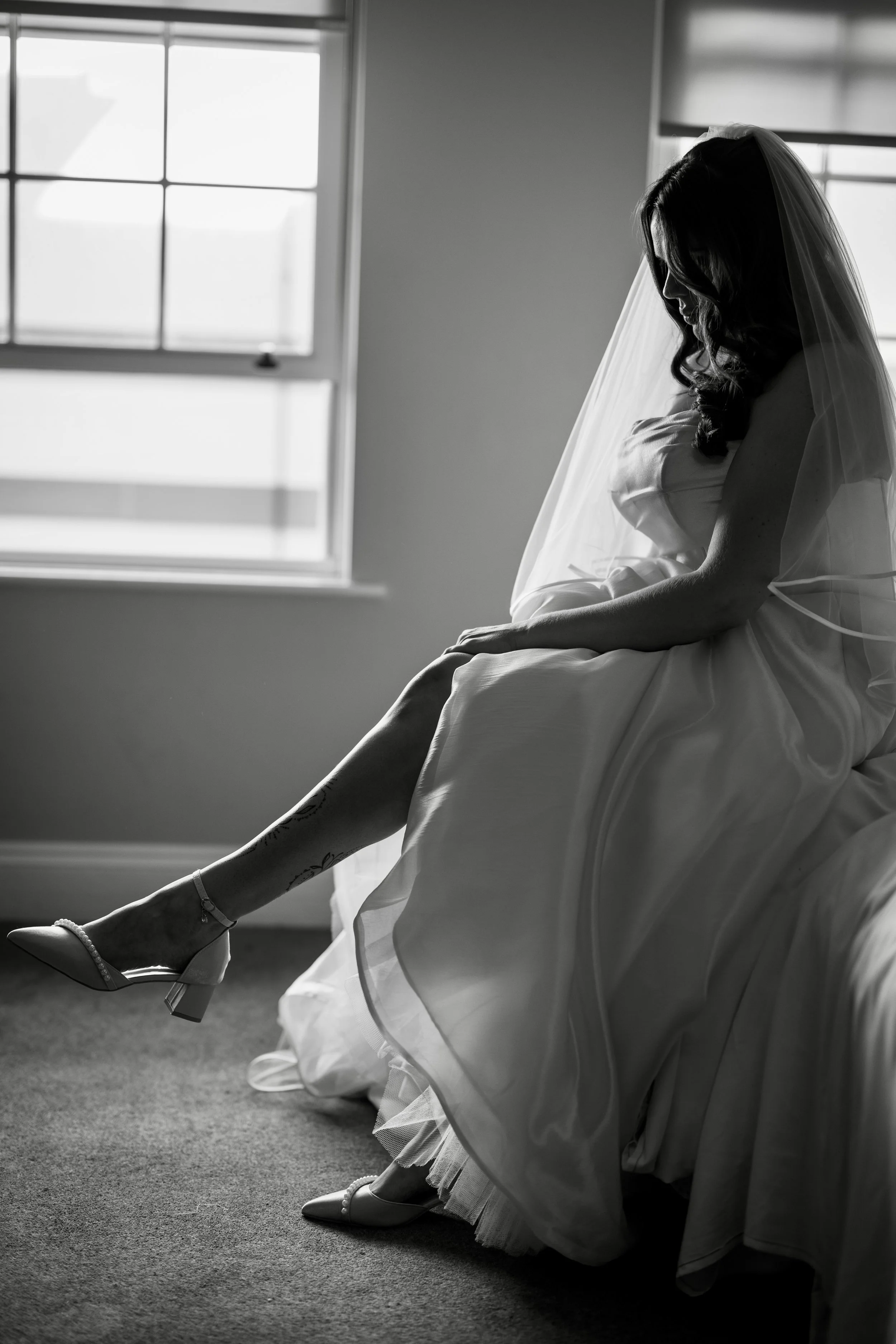 A bride sitting by a window, looking down, wearing a wedding dress, veil, and high heels in a black-and-white photo.