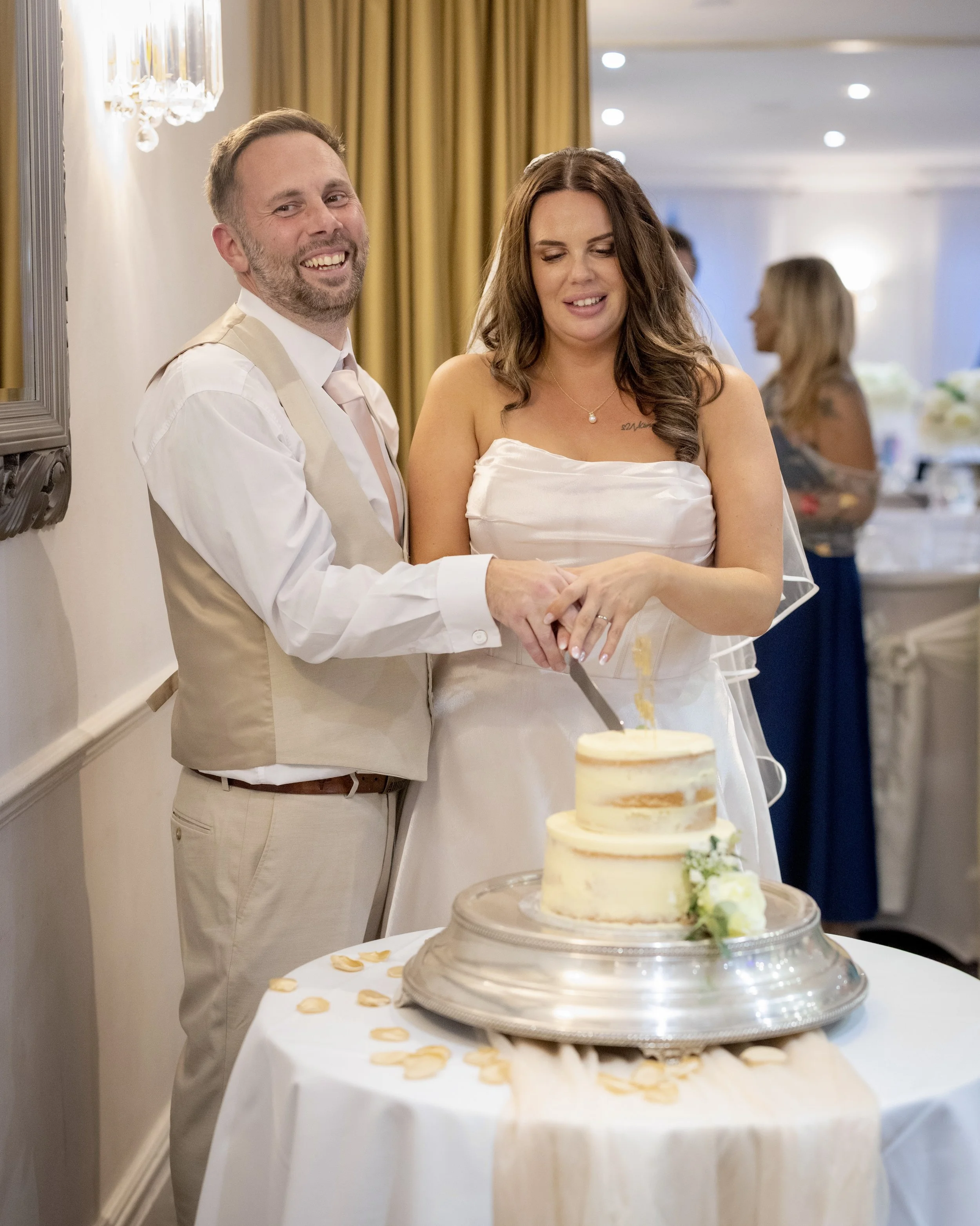 A newlywed couple at their wedding reception cutting a three-tier wedding cake, with friends and family in the background.