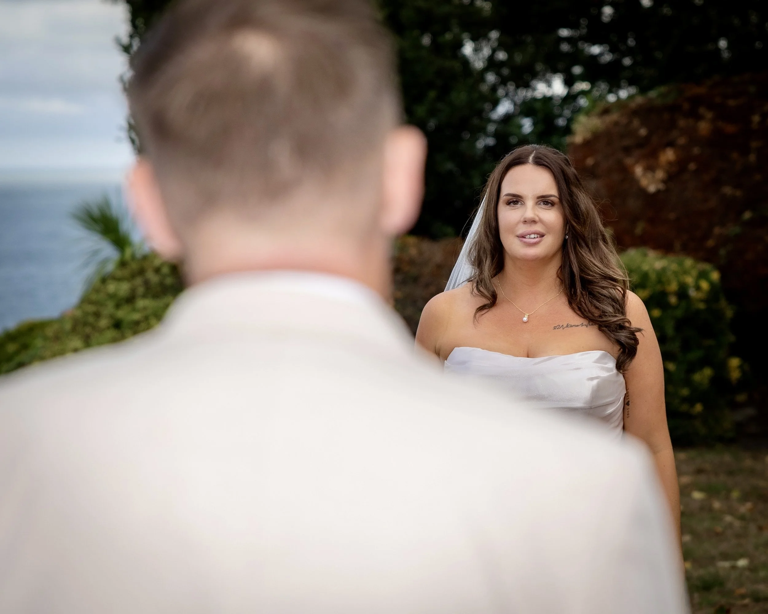 A bride with long brown hair and a tattoo on her shoulder, wearing a white strapless dress, stands outdoors with trees and a blurred background, looking at a man in a white suit with a close-cropped haircut.
