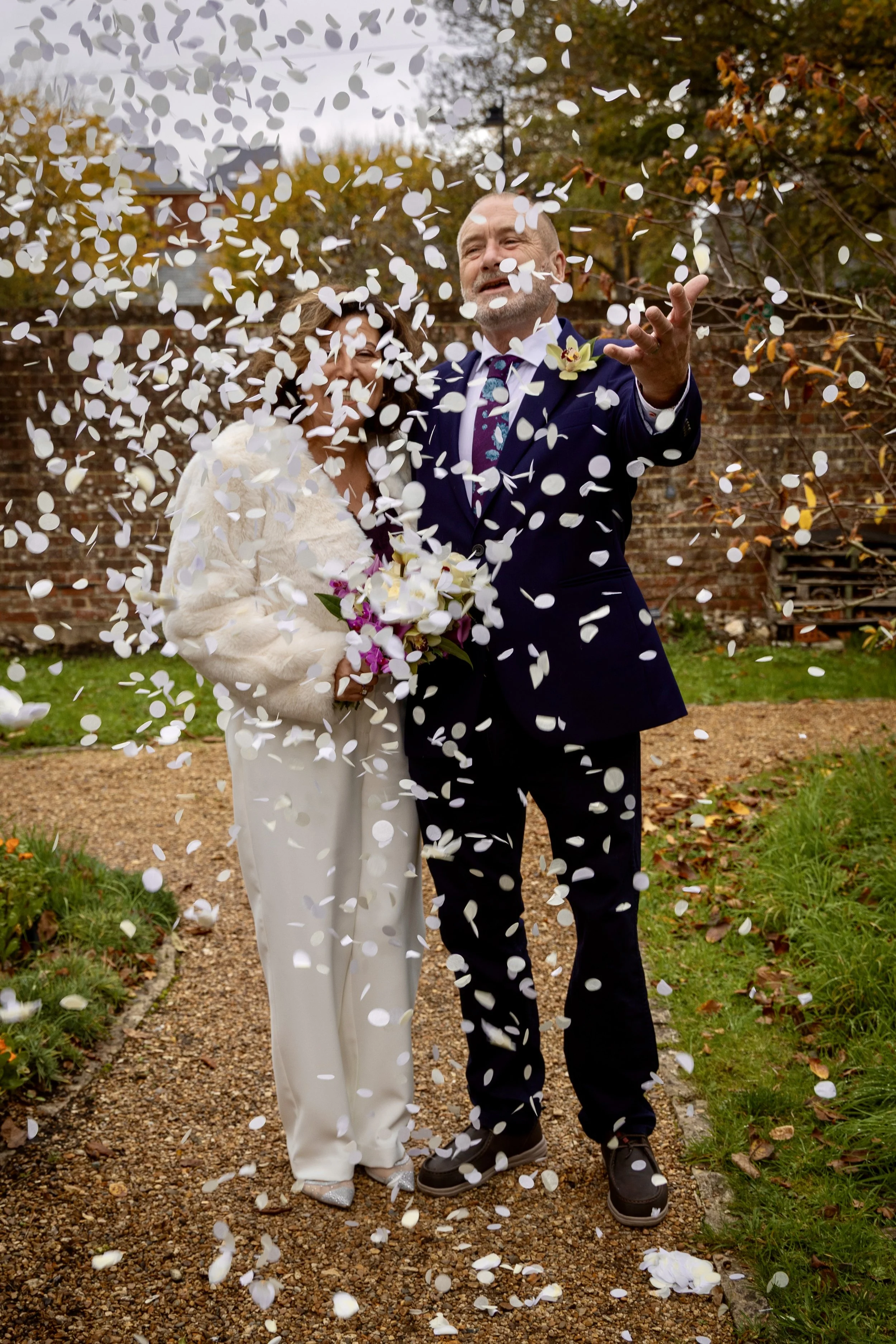 A bride and groom celebrating their wedding outdoors with confetti falling around them, standing on a gravel path surrounded by trees with fall foliage.