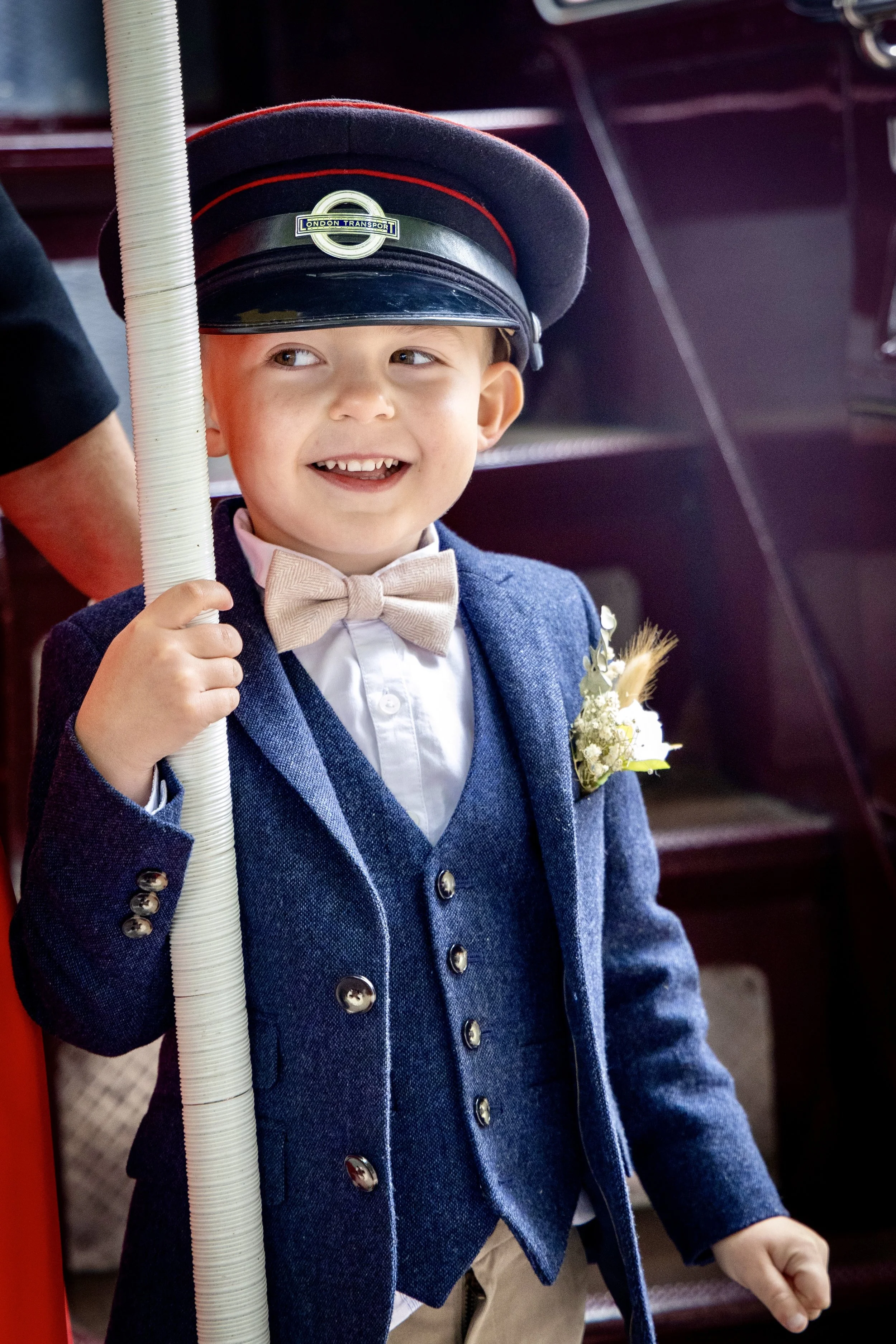 Young boy dressed in a blue blazer with a boutonniere, wearing a hat that says 'London Transport', holding a white pipe, smiling and looking to the side.