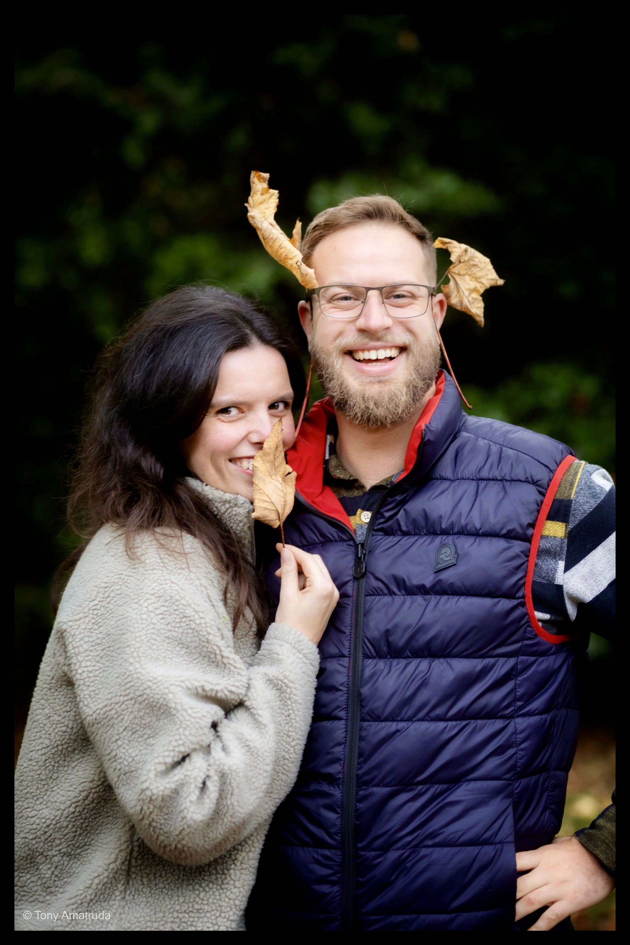 A smiling couple outdoors with fall leaves, green background, there's a woman with long brown hair and a beige jacket, and a man with glasses, beard, and a blue vest, as leaves are on their heads and in their hands.