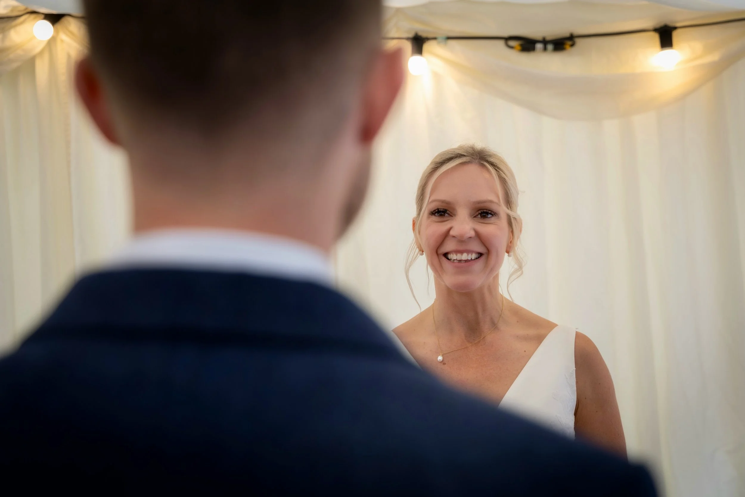 A woman in a white dress smiling, seen through the blurred shoulder and head of a man in a suit, at what appears to be a wedding or celebration under a tent with string lights.