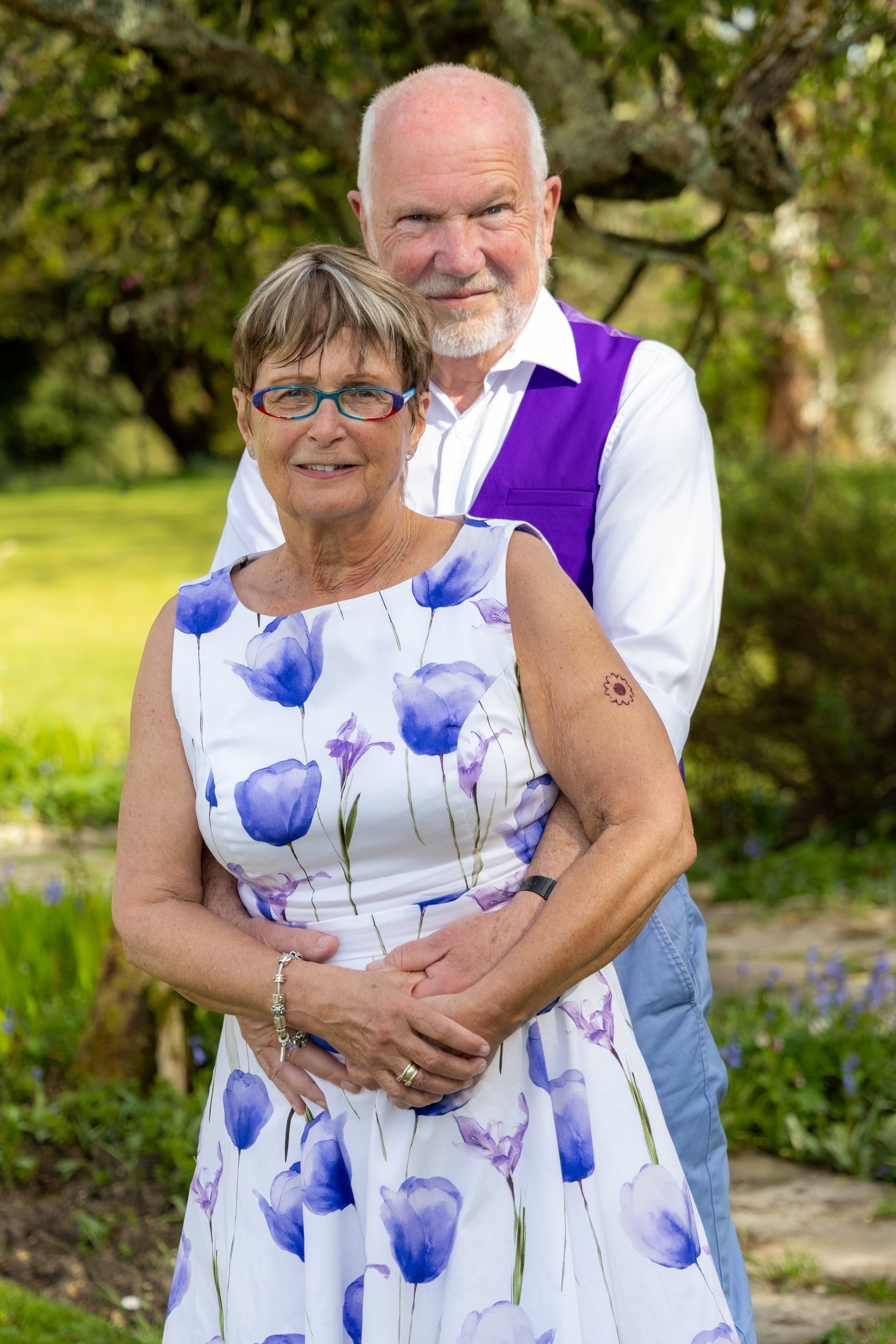 A smiling elderly woman wearing a white dress with blue floral pattern, standing in front of a man with a bald head and white beard, outside in a green park with trees and bushes.