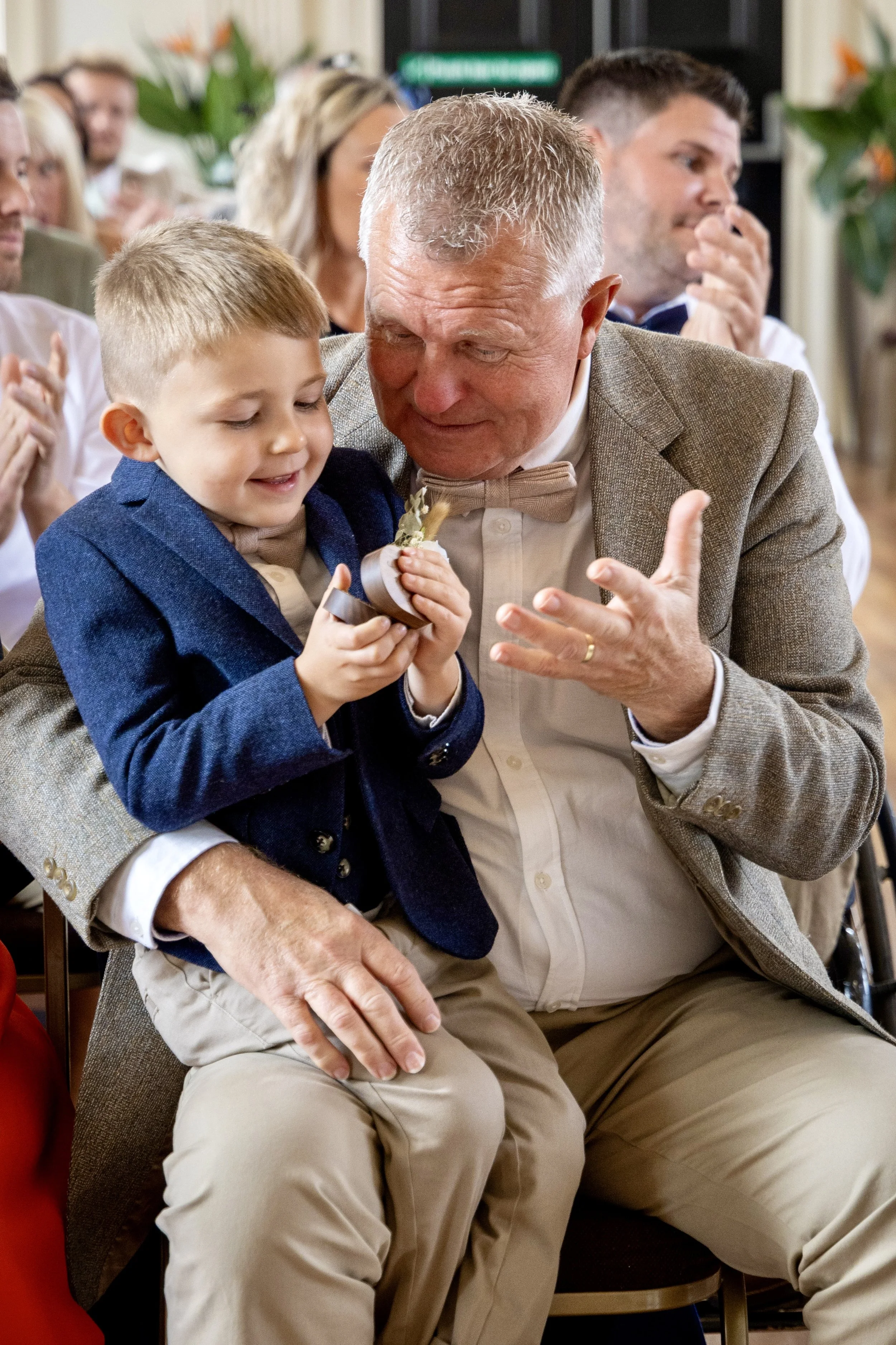An elderly man and a young boy, sitting close together at a formal gathering, sharing a moment of joy. The boy is holding a medal and smiling, while the man is leaning in affectionately, both wearing dress shirts and bow ties.