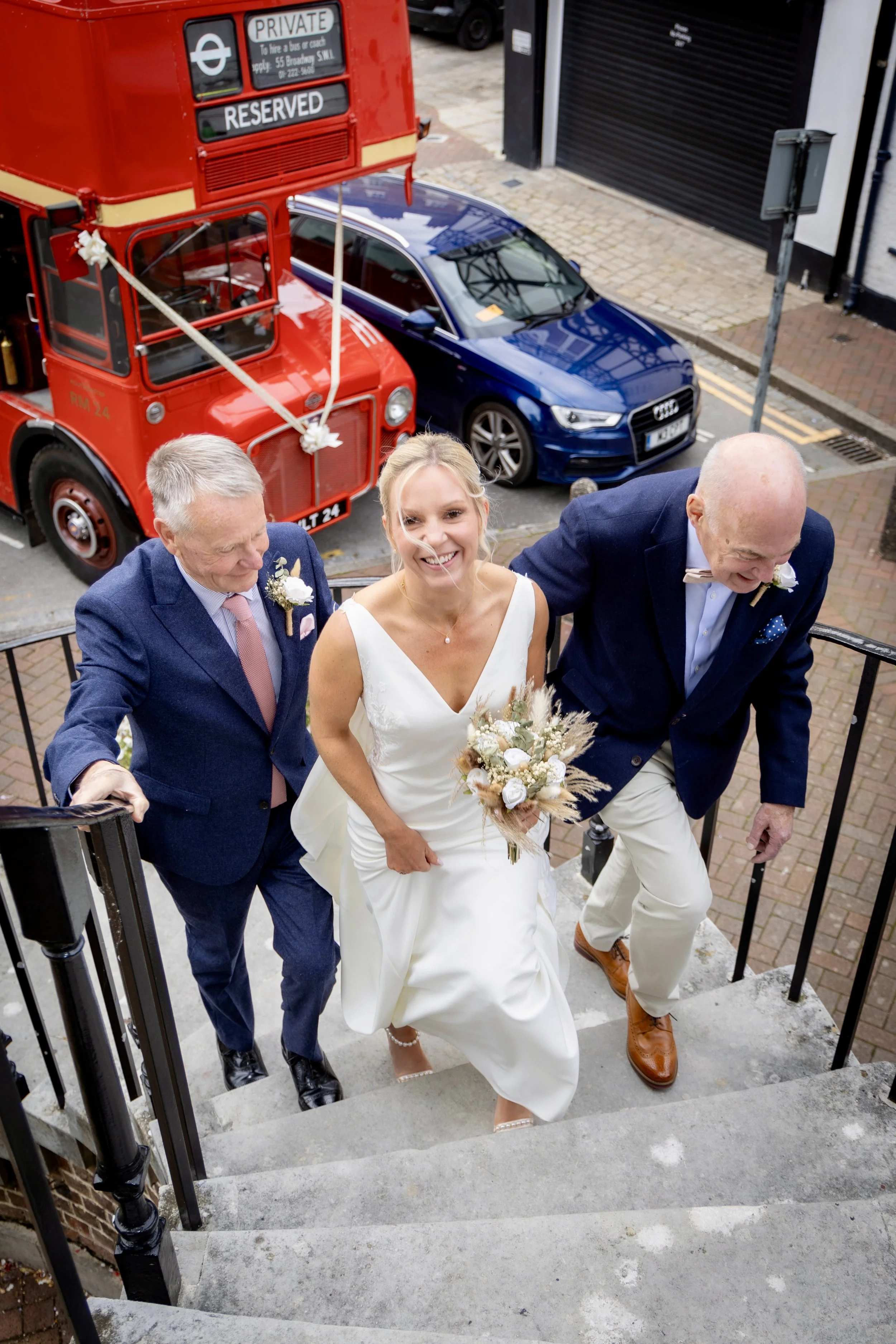A bride and two men walking up outdoor stairs during a wedding celebration. The bride is holding a bouquet of white and beige flowers and is smiling. The men are dressed in suits, one in navy and the other in dark blue, with boutonnières. In the back