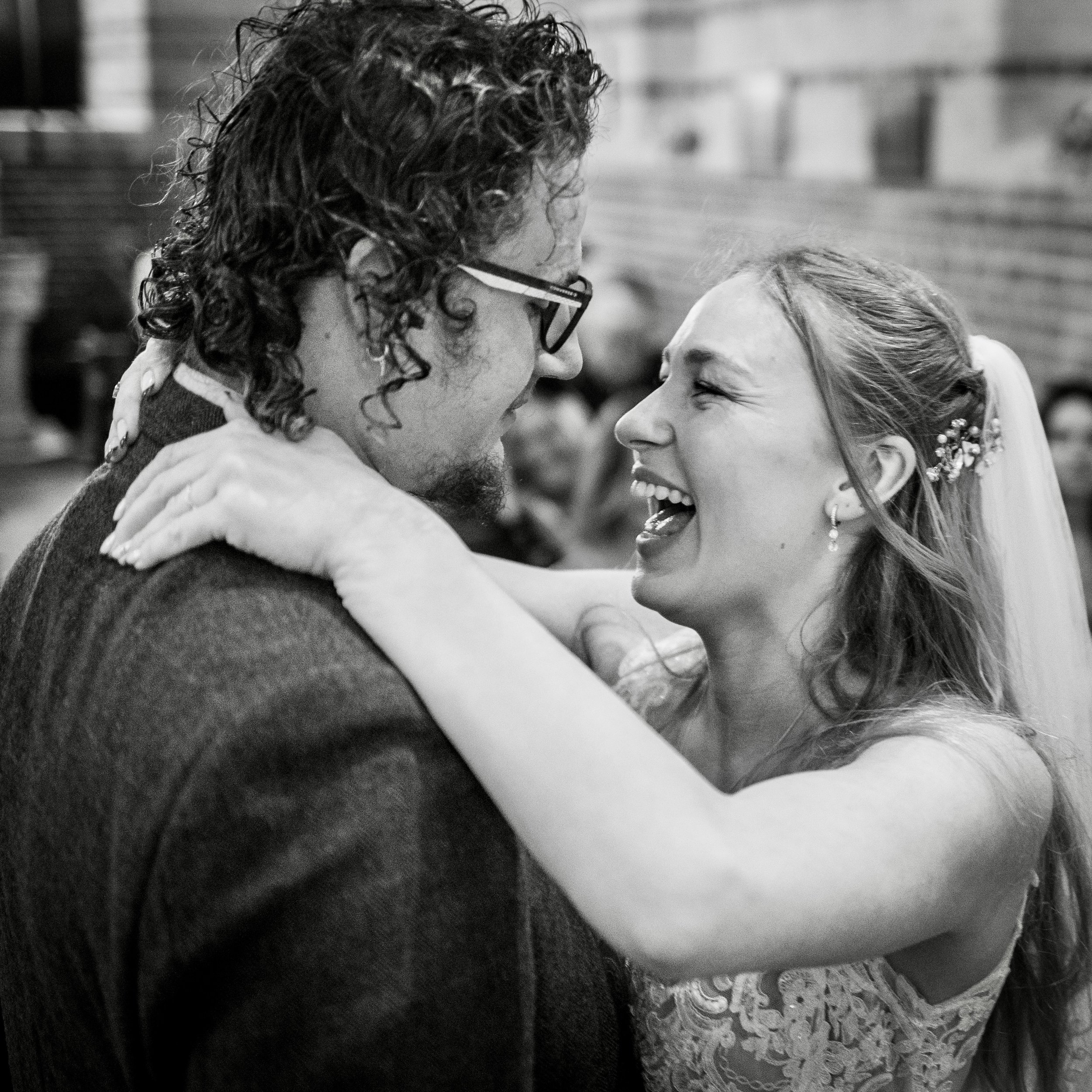 A black and white close-up photo of a man and woman smiling and looking at each other, possibly at a wedding or special event, with blurred background.