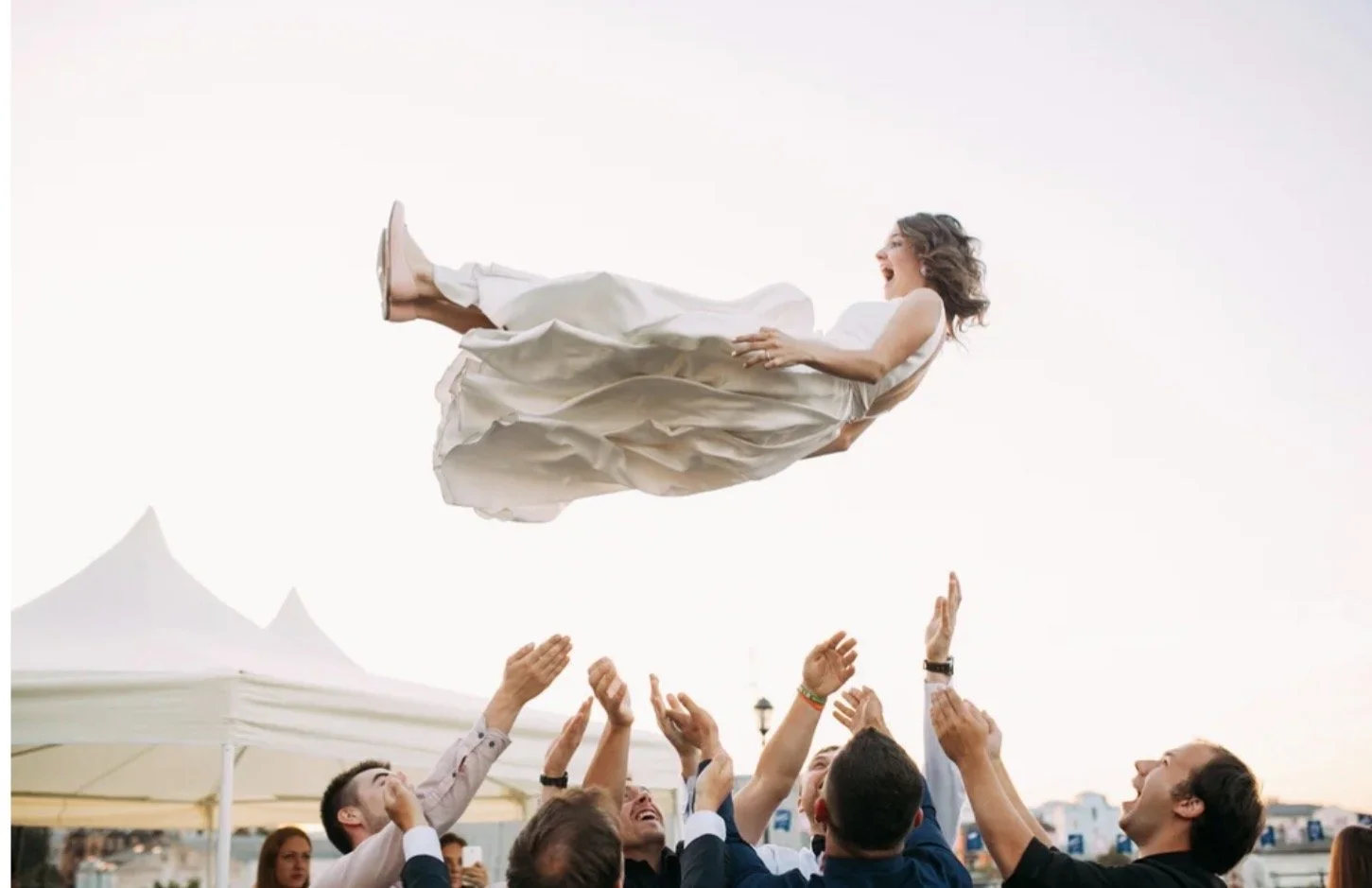 Men and women celebrating outdoors as a woman in a white dress is lifted into the air, laughing, against a bright sky.