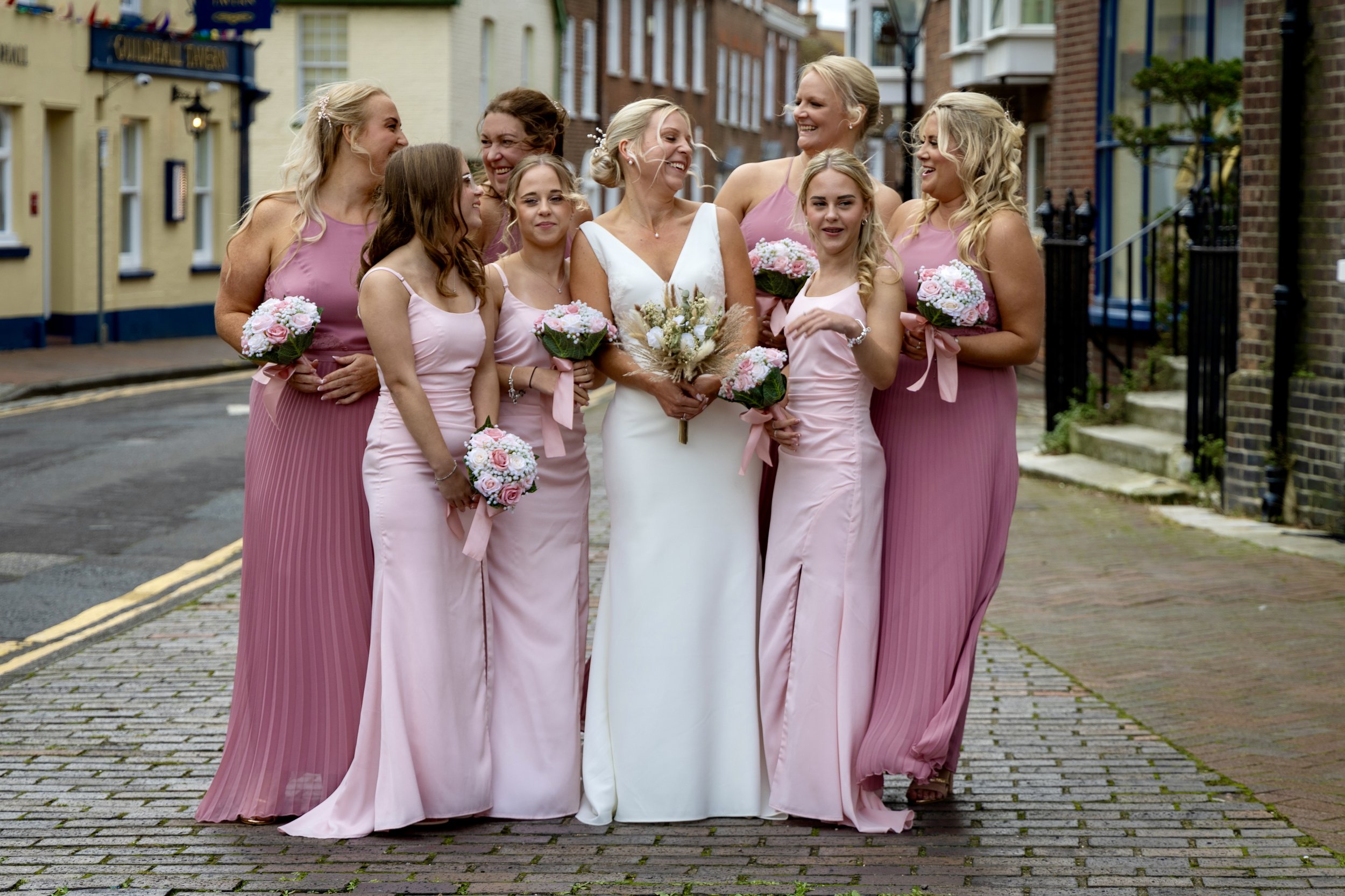 A bride in a white wedding dress surrounded by eight women in pink dresses, all holding bouquets of pink and white flowers, standing on a cobblestone street in an urban area.