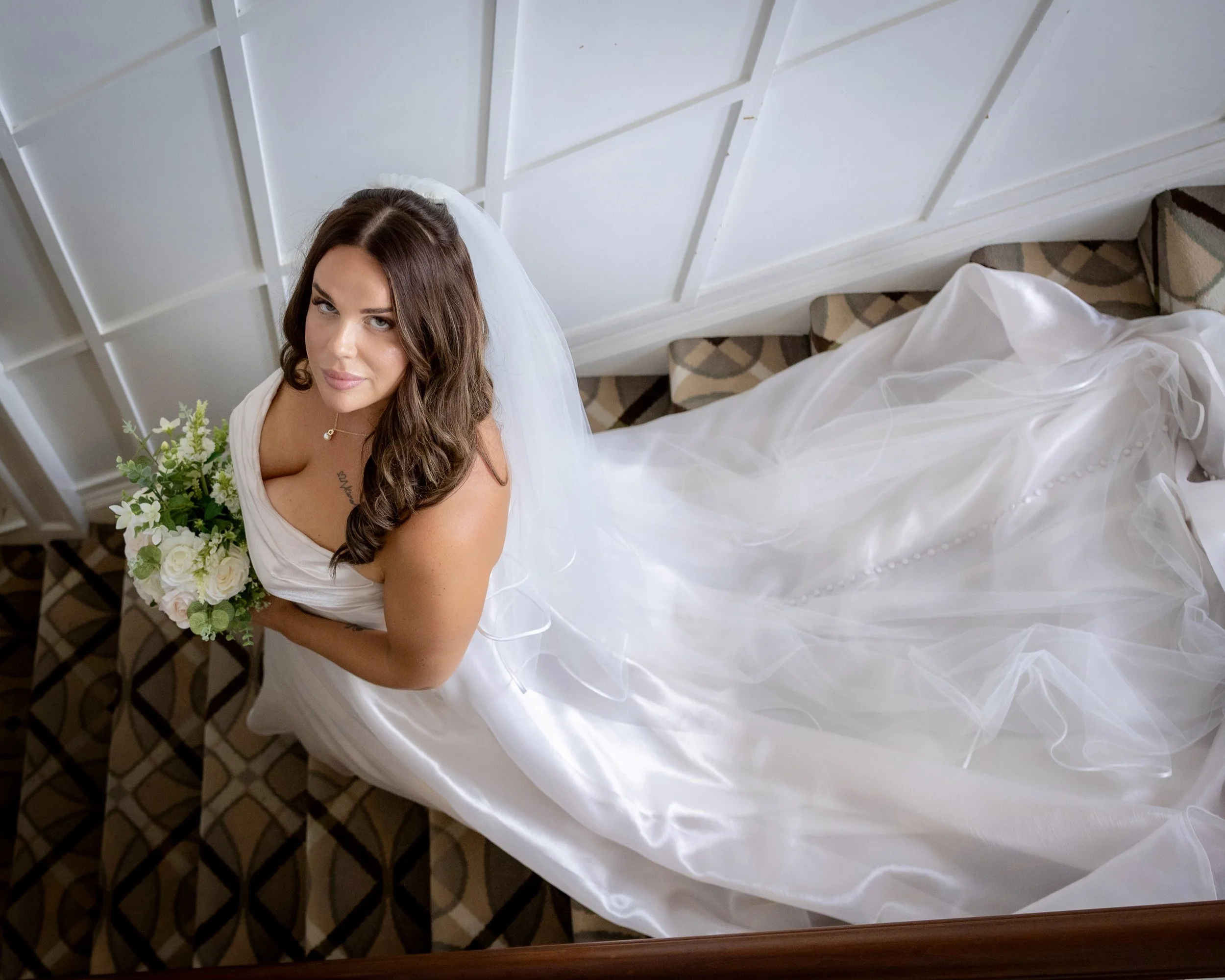 A woman dressed in a white gown holding a bouquet of flowers, sitting on a staircase with a geometric patterned carpet, photographed from above.