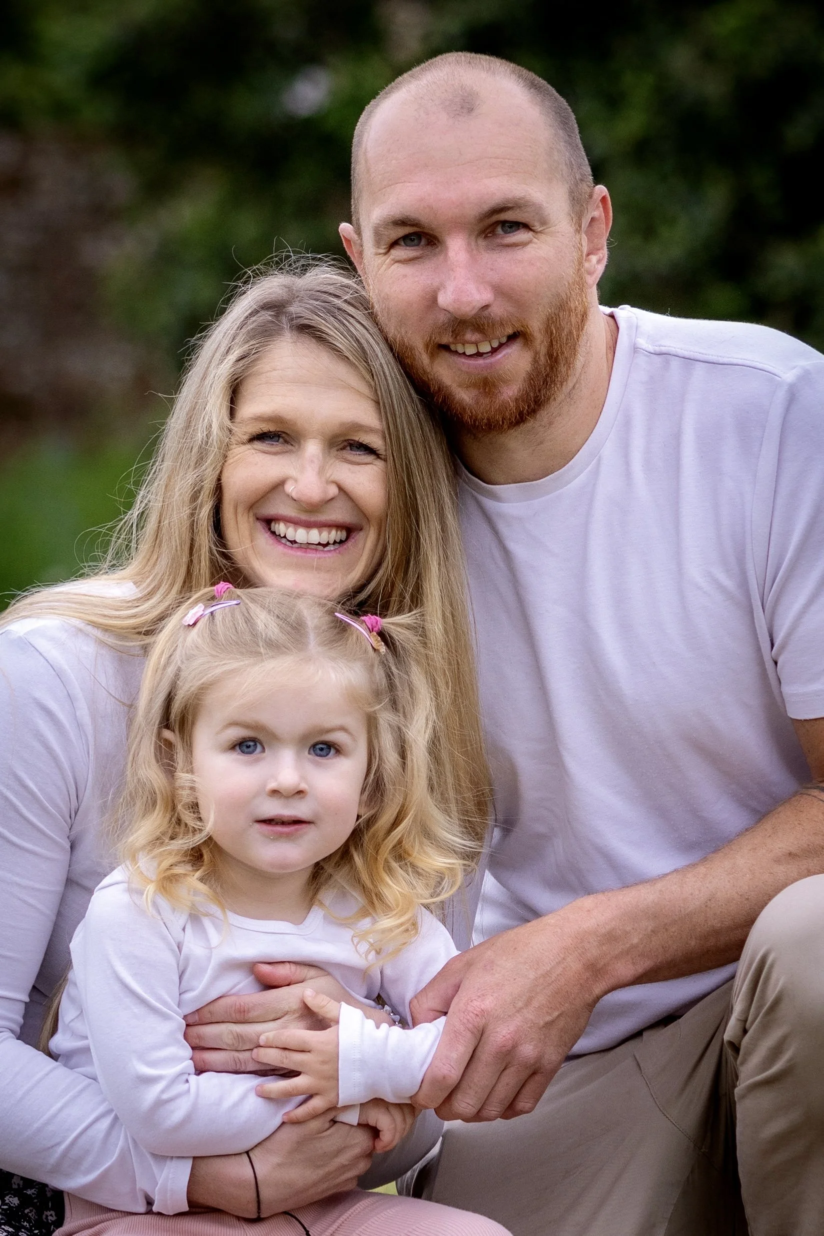 A family portrait of a man, woman, and young girl outdoors against a blurred green background. The woman has long blonde hair, the man has a red beard and is bald, and the girl has curly blonde hair tied with pink hair ties. They are smiling and close together.