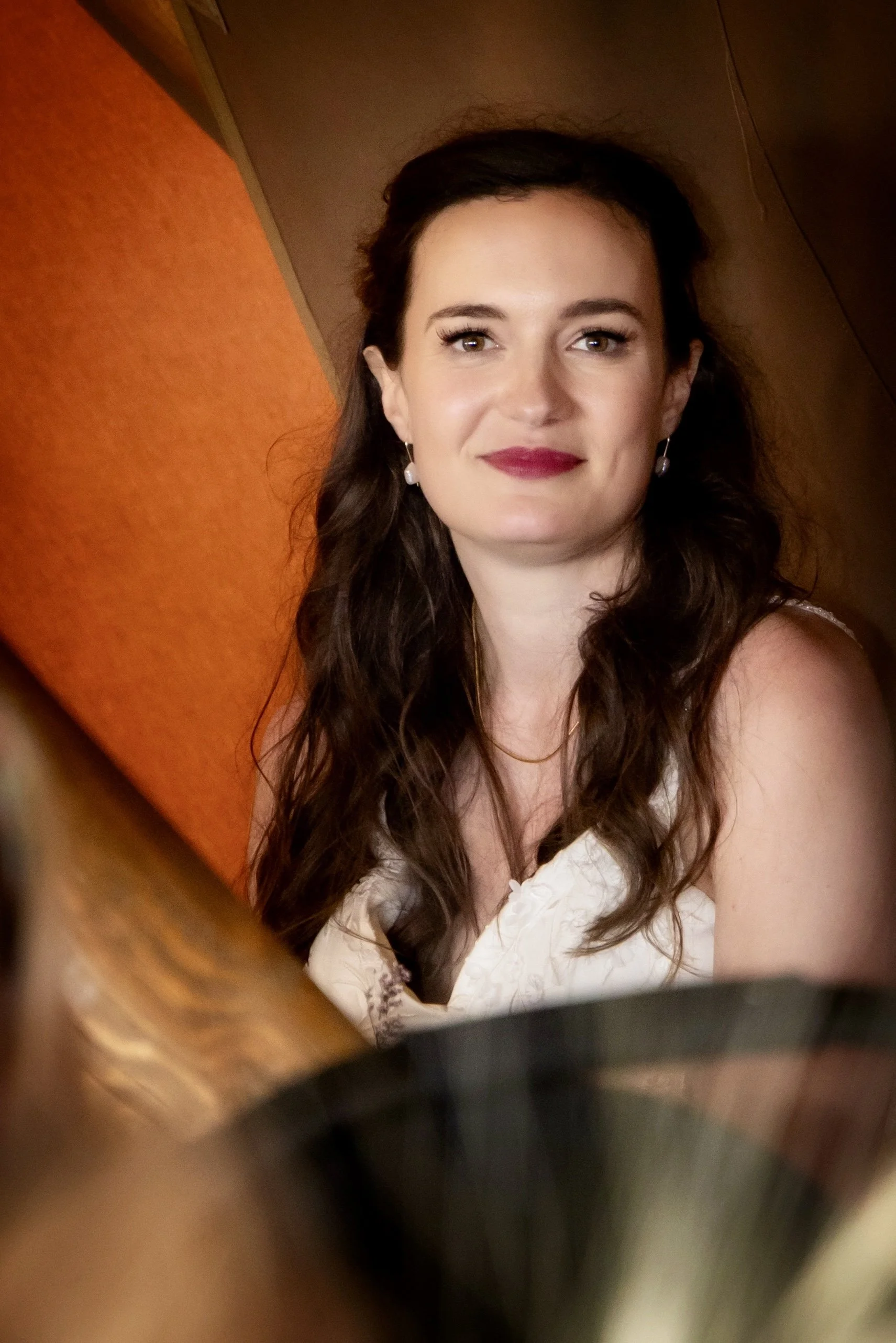 A woman with long, wavy brown hair, makeup, and jewelry sitting at a table with an orange wall behind her.