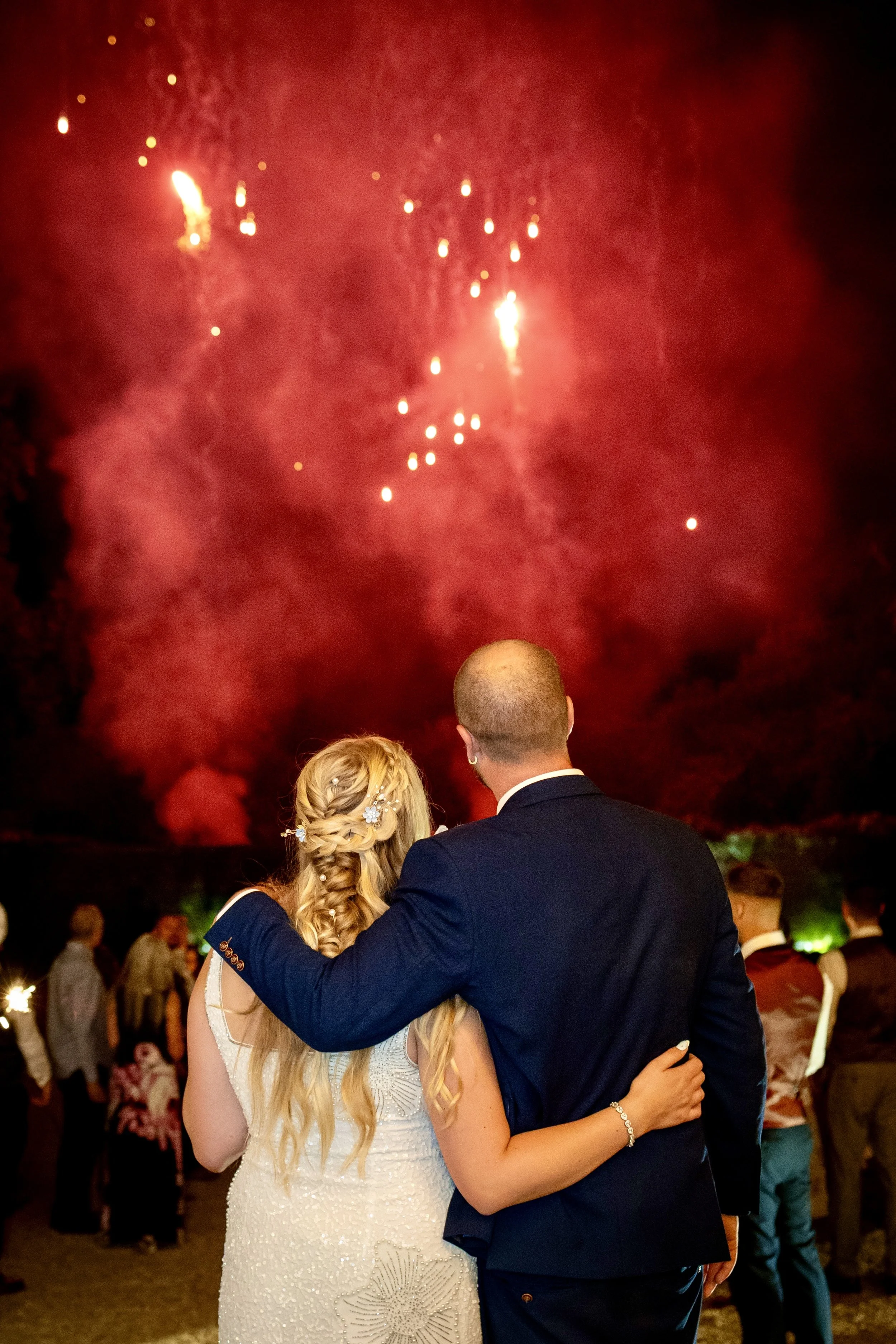 A couple in wedding attire watching fireworks at night, with a group of people in the background.