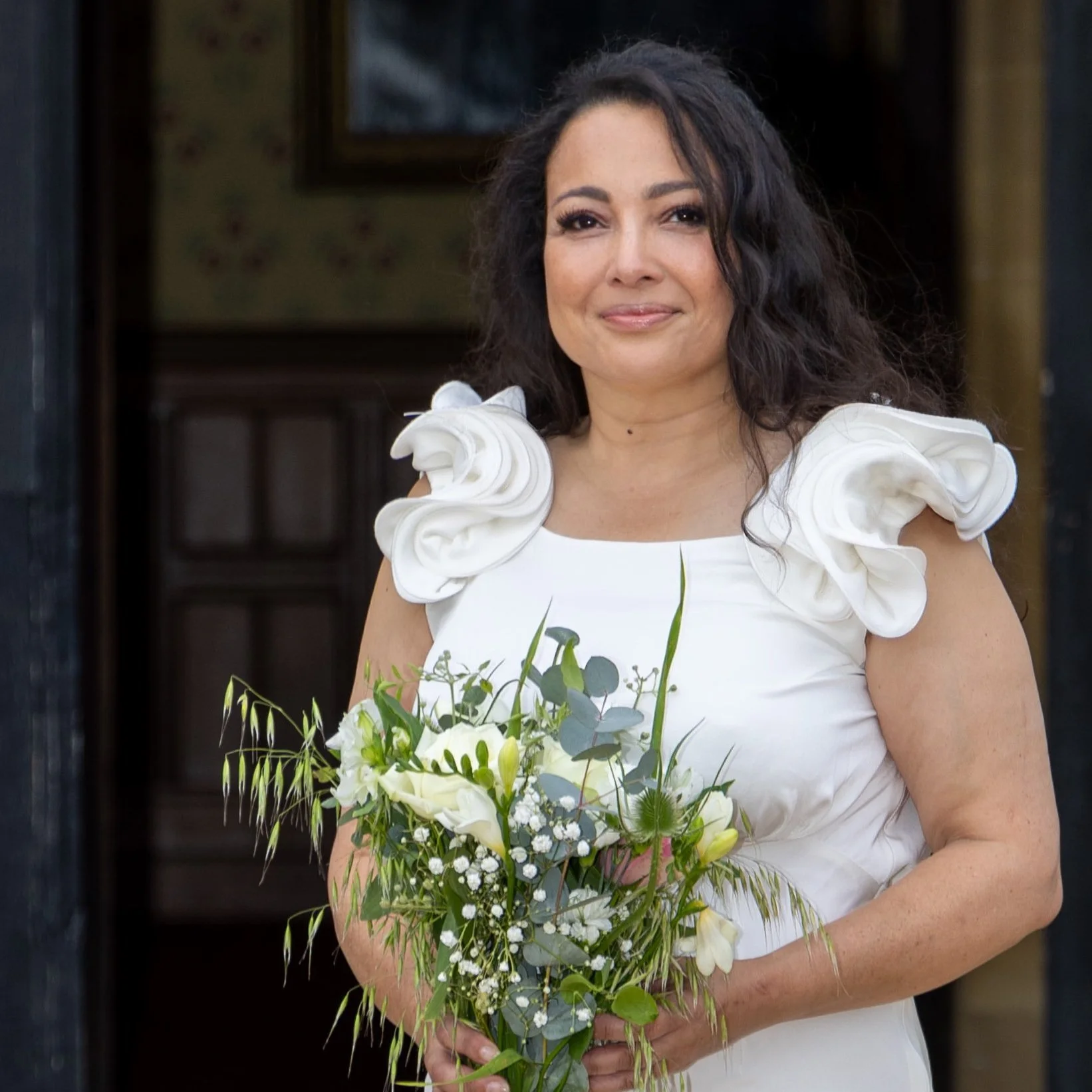 A woman dressed in a white dress with ruffled shoulder details holding a bouquet of white flowers and greenery at what appears to be a wedding or special event.