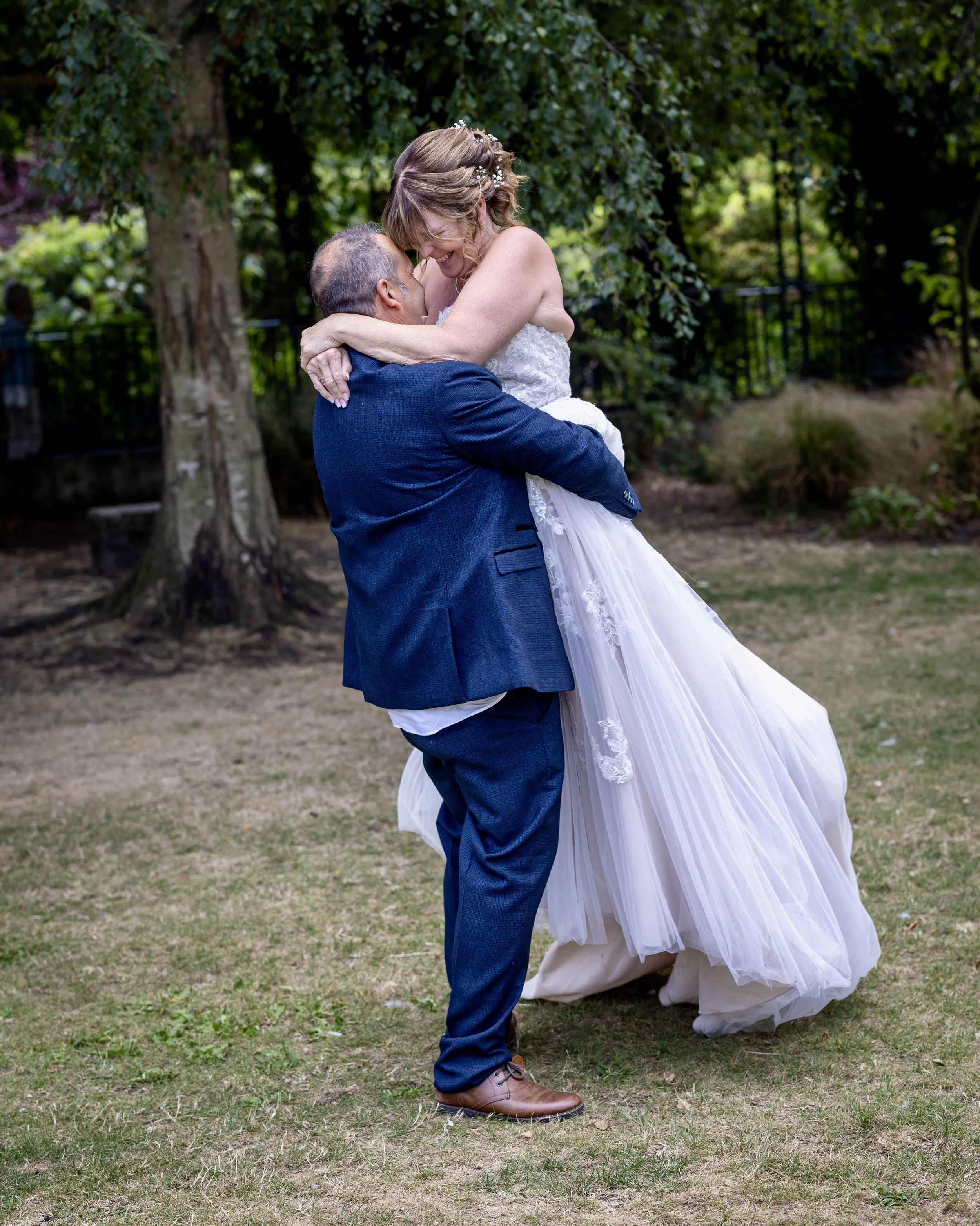 A woman in a wedding dress is being lifted by a man in a suit outside in a garden. They are embracing and smiling, with trees and greenery in the background.