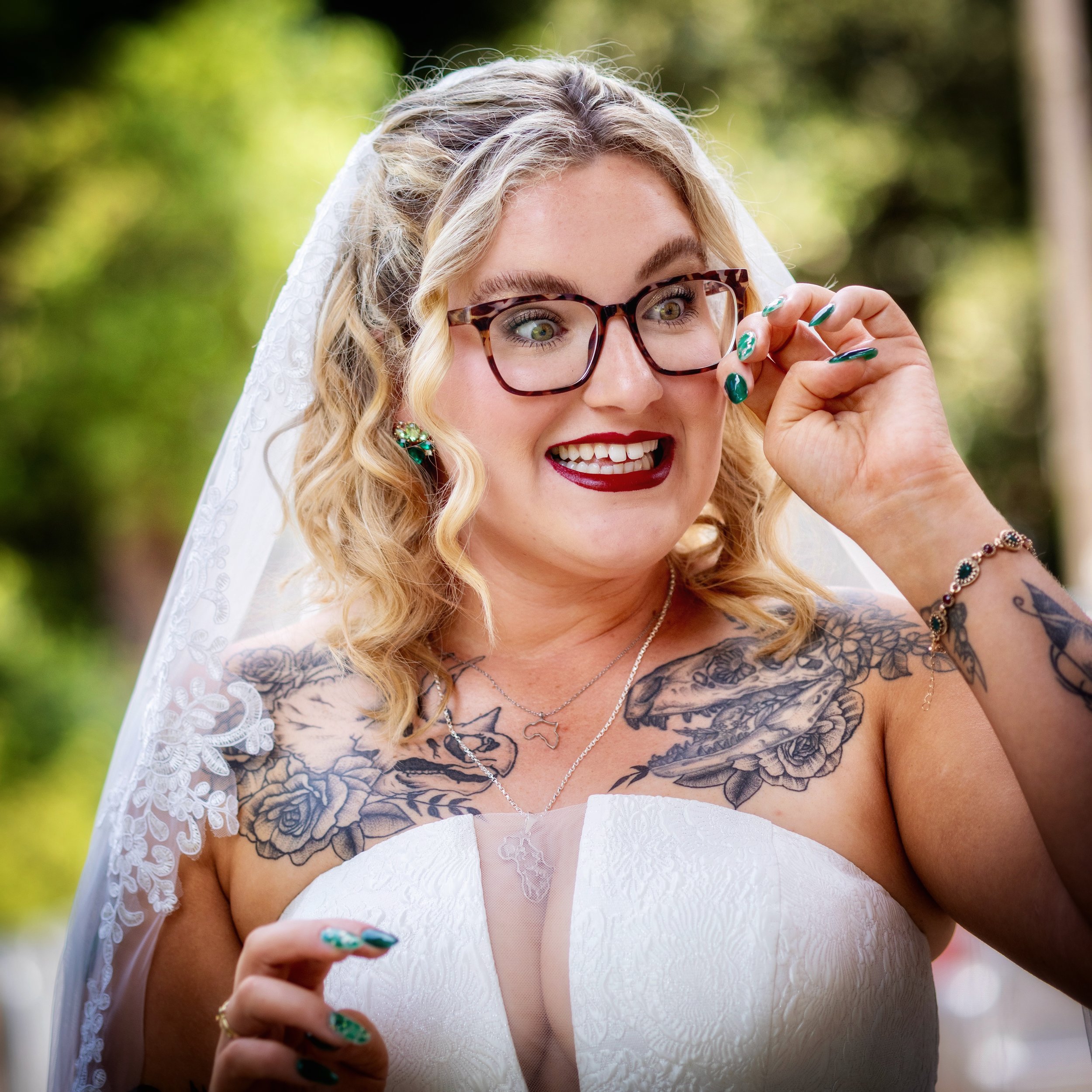 A woman with blonde curly hair, glasses, and tattoos on her shoulders dressed in a wedding gown and veil, smiling and adjusting her glasses outdoors.