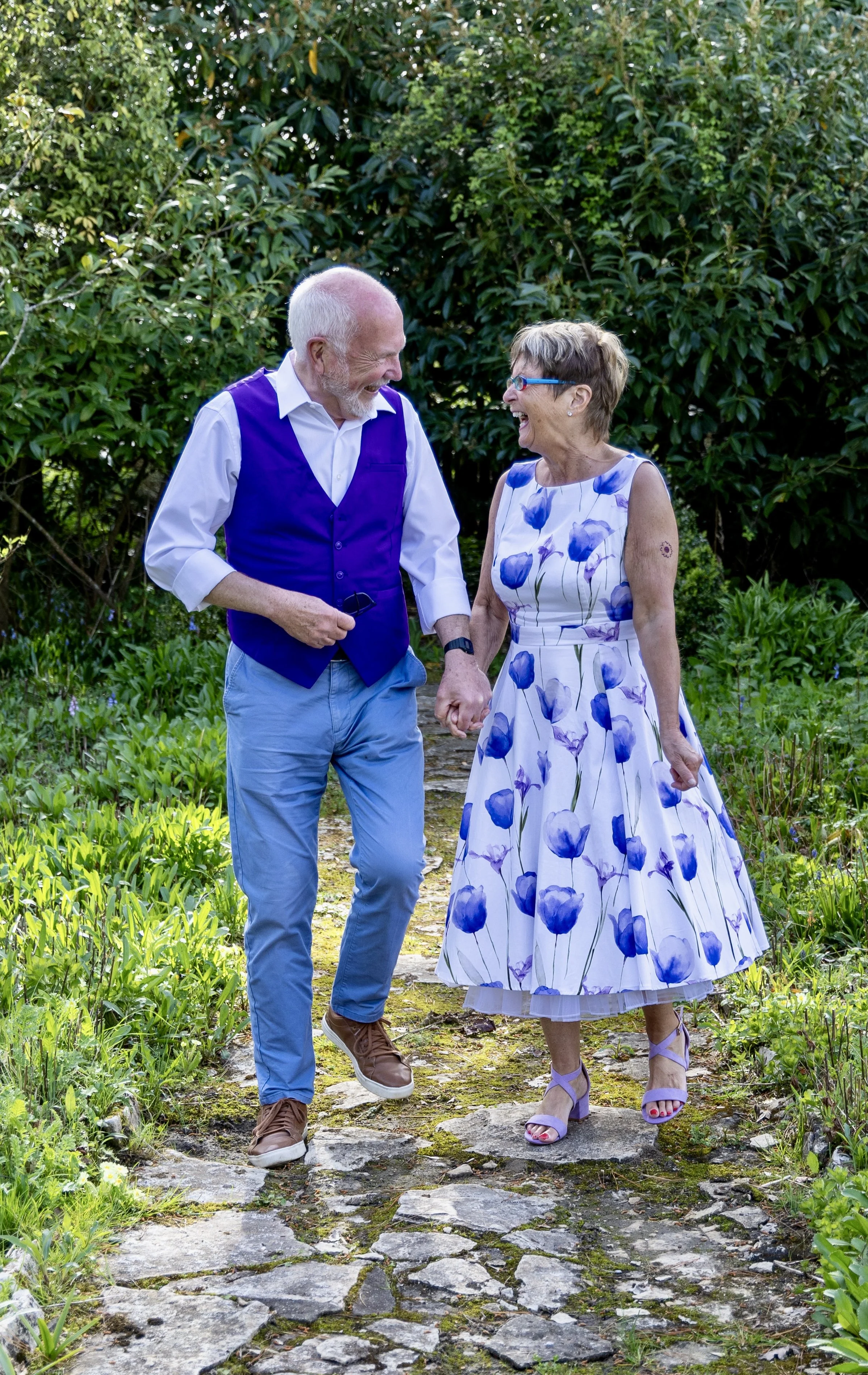 An elderly couple holding hands and laughing, walking along a stone path in a lush garden. The man is wearing a blue vest, white shirt, and light blue pants, while the woman is in a white dress with purple flower patterns and purple high heels. They 