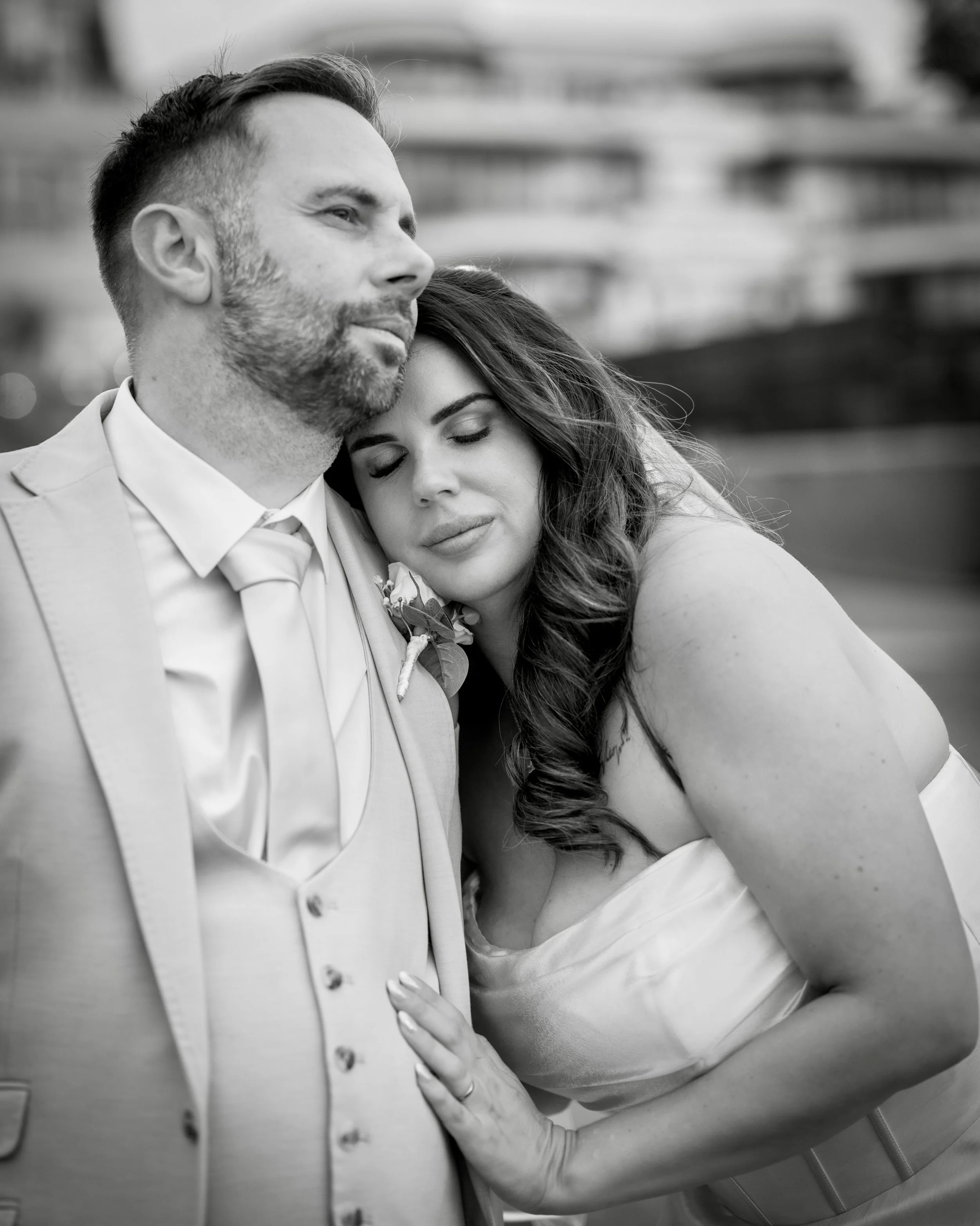 A black and white photo of a happy couple with their eyes closed, embracing each other. The man is wearing a formal suit with a tie, and the woman has long, wavy hair and is wearing a dress or wedding gown. The woman’s arm is wrapped around the man, 