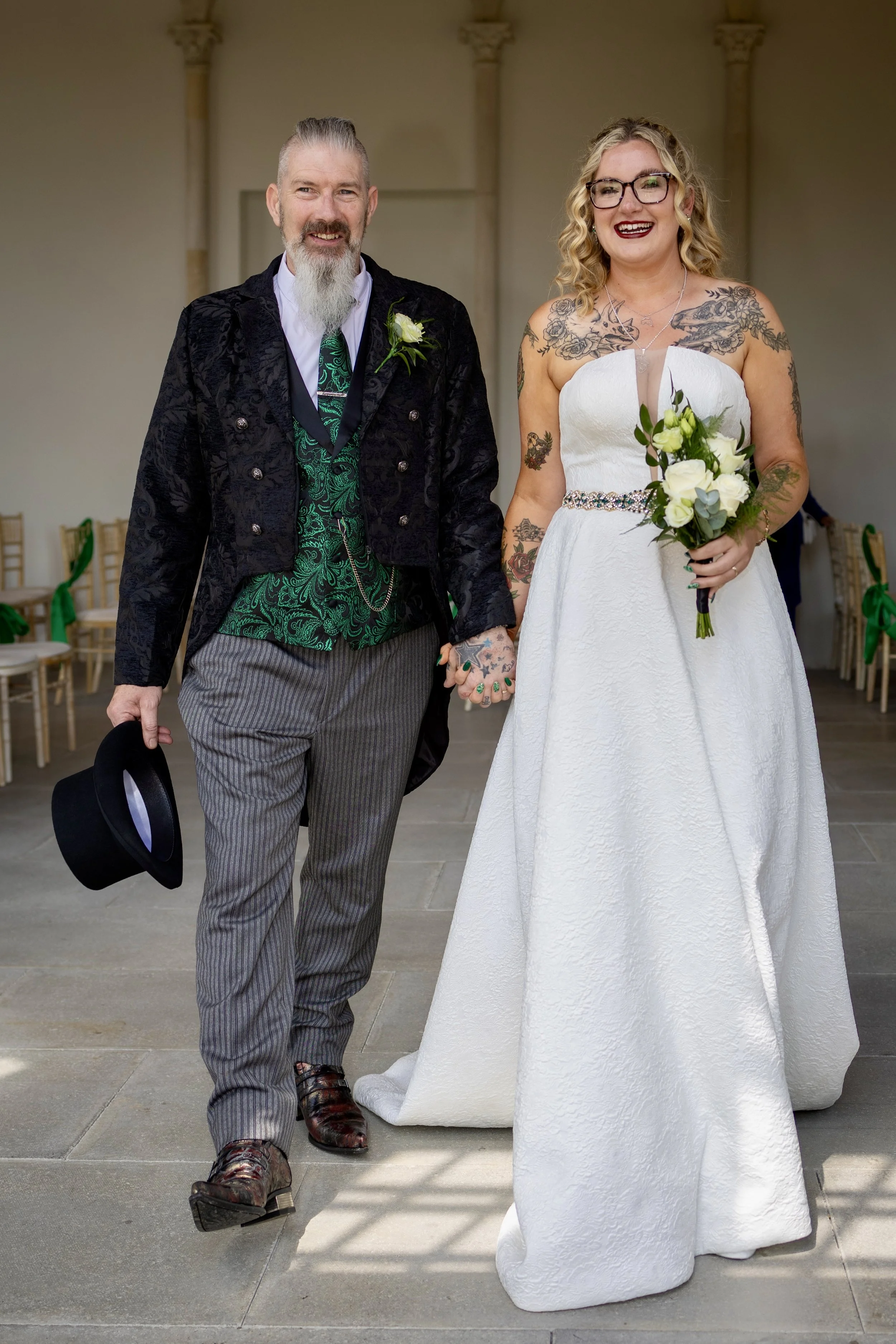 A man and woman holding hands, dressed in wedding attire, standing indoors. The man is wearing a black velvet jacket, striped pants, and patterned shoes, holding a black hat. The woman is in a strapless white wedding gown with a bejeweled belt, and h