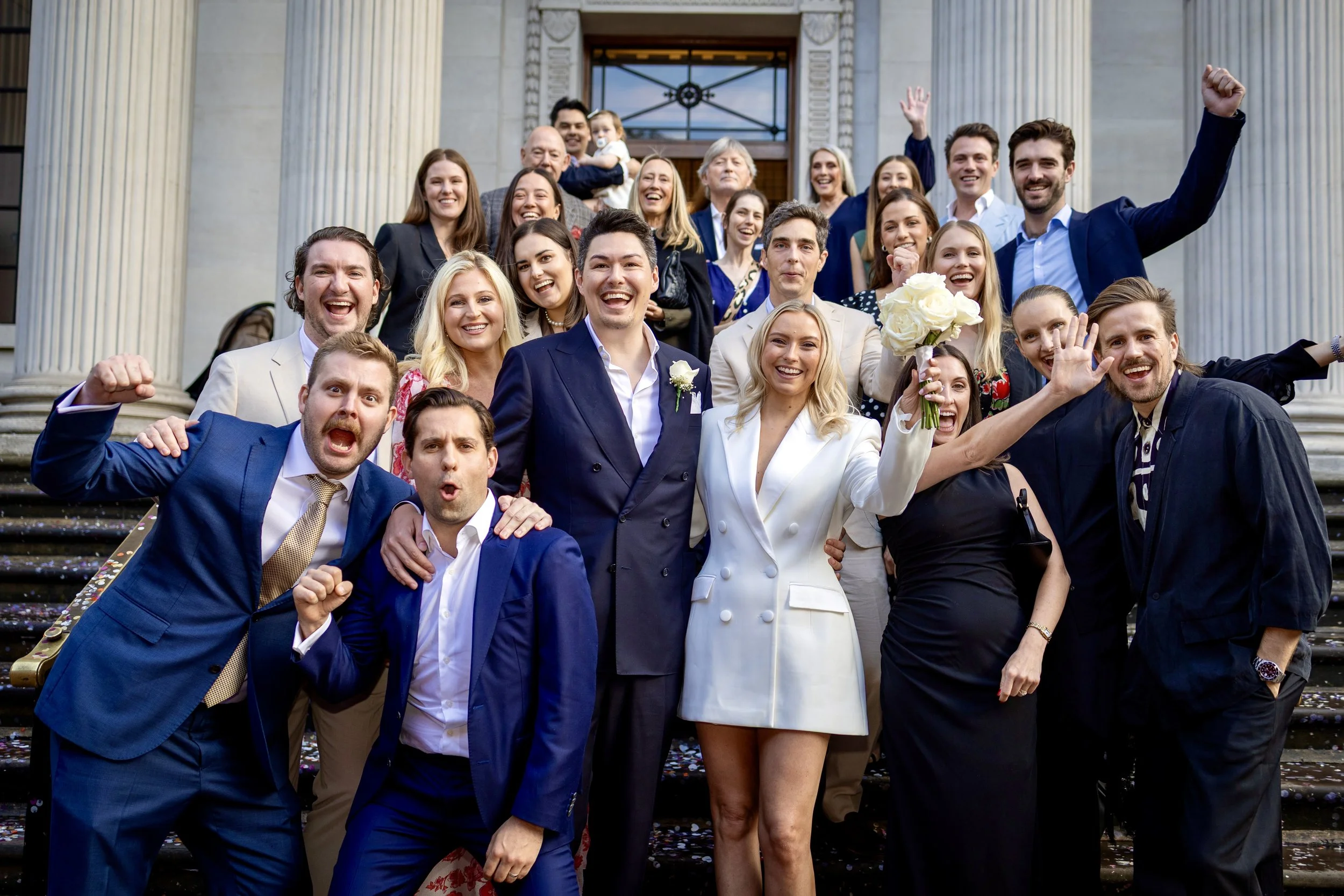 Group of wedding guests celebrating on the steps of a building with columns, some making cheering gestures, smiling, and dressed in formal attire.