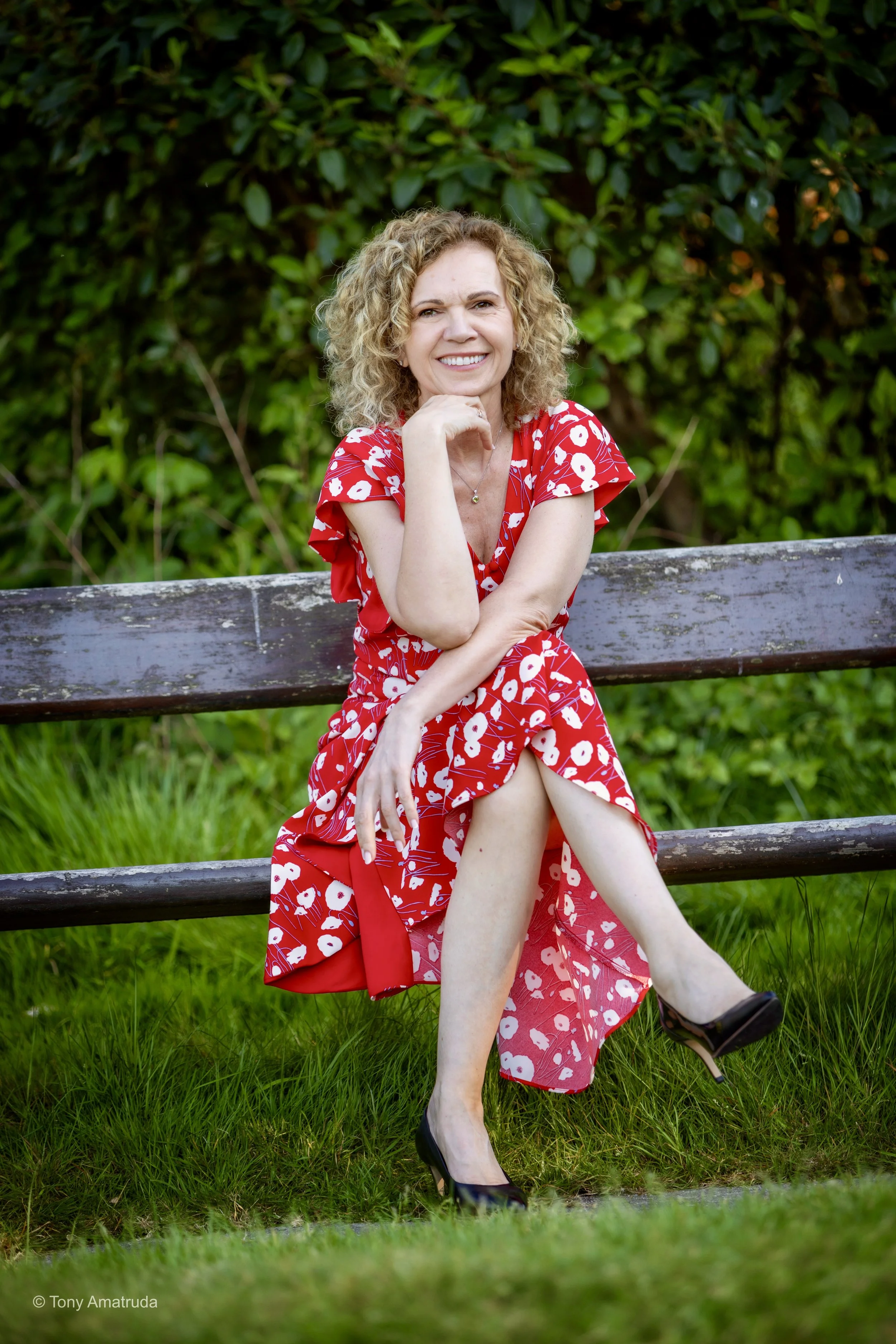 Woman with curly blonde hair wearing a red dress with white flower pattern, sitting on a wooden bench outdoors with greenery background, smiling and posing with one hand on her chin
