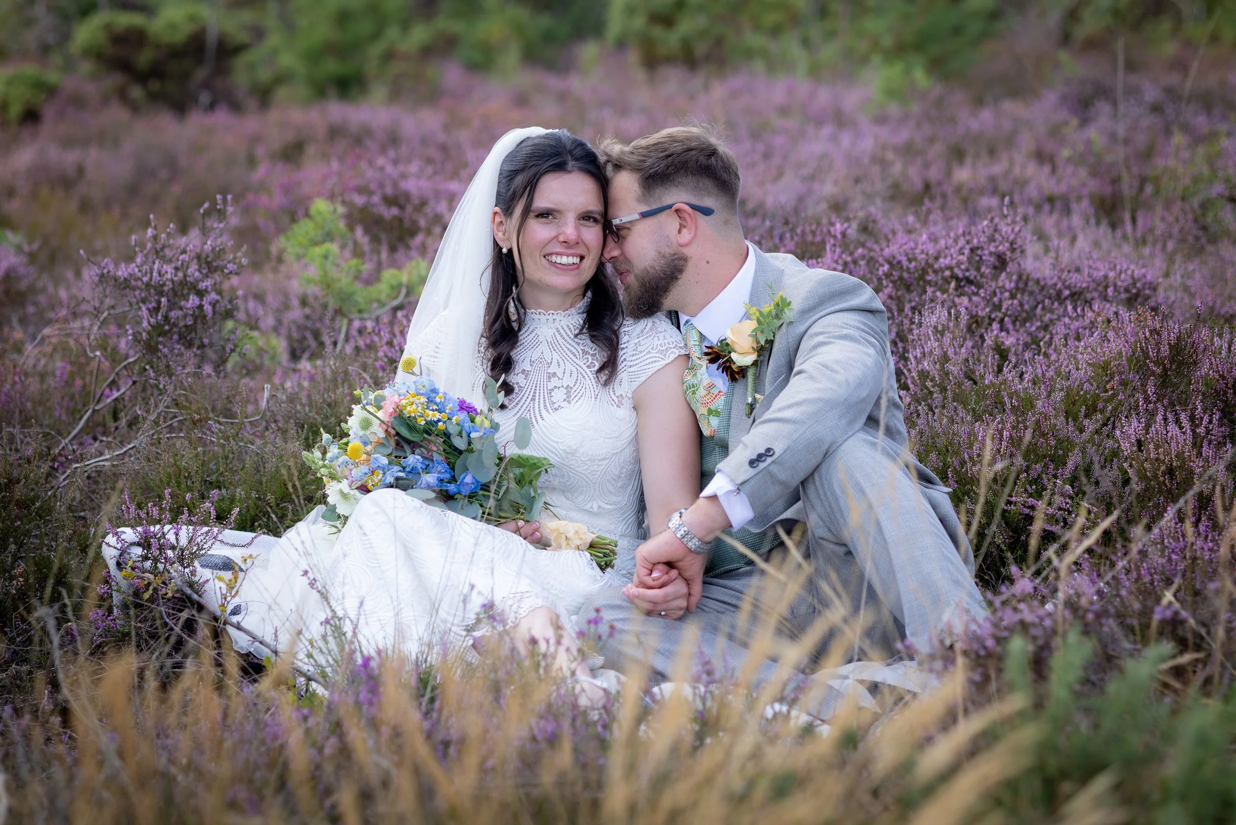 A bride and groom sitting in a purple heather field during their wedding, with the bride holding a colorful bouquet and smiling at the camera while the groom whispers to her.