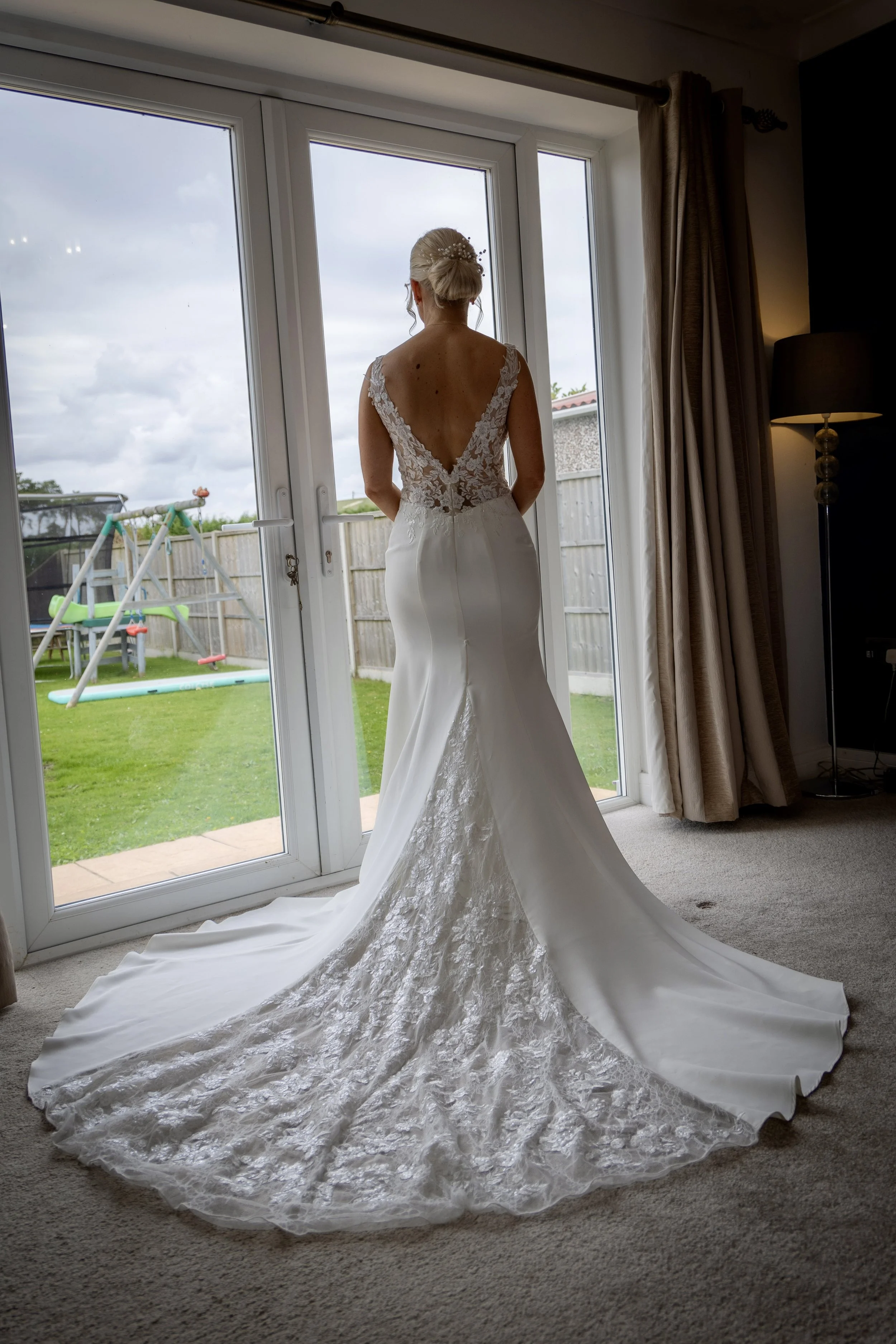 A bride standing inside near large glass doors, wearing a white wedding gown with lace details on the back and a long train. She is looking outside at a backyard with a playground and a cloudy sky.