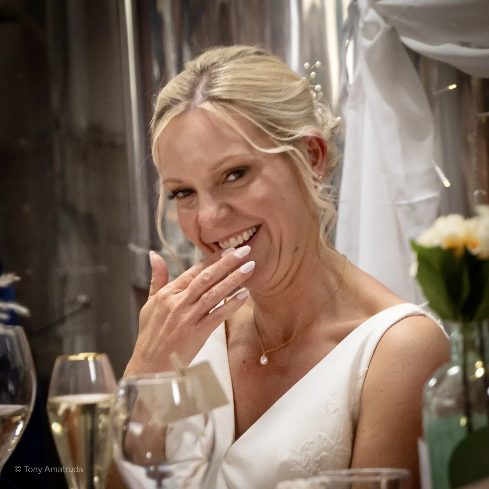 A smiling woman in a white dress, with blonde hair, at a celebration or formal event, with glasses and flowers on the table in front of her.
