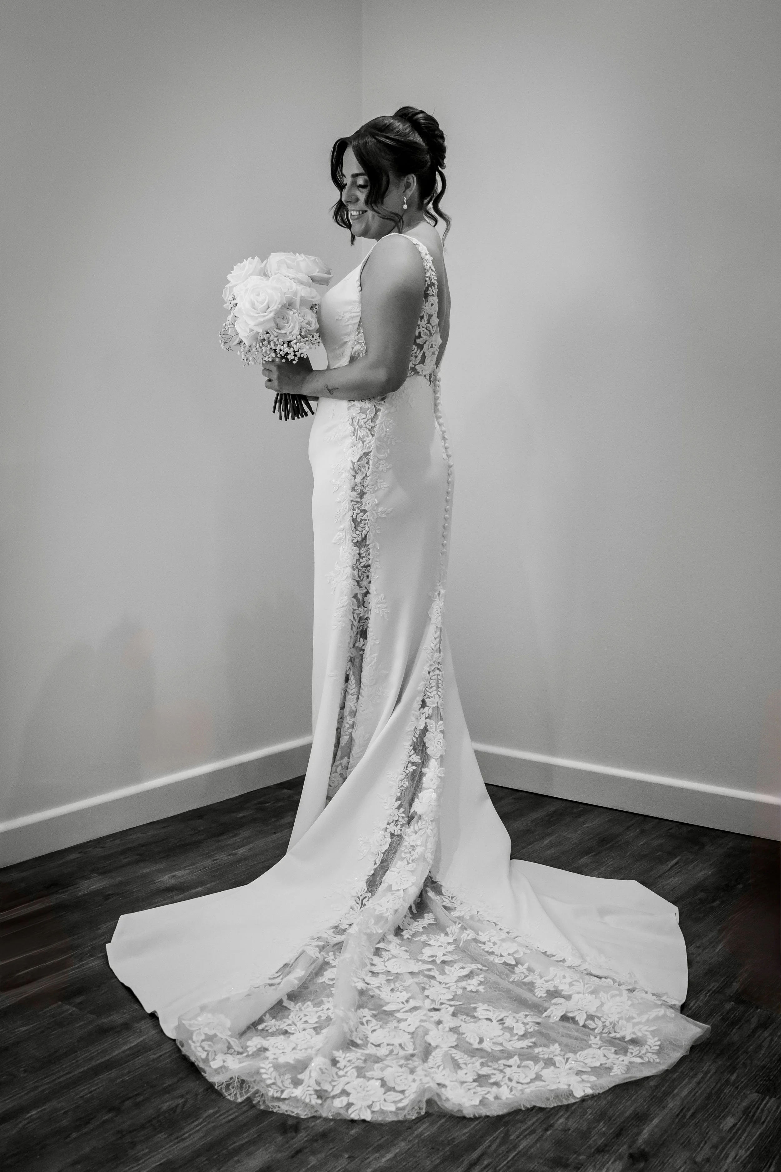 A bride in a white wedding gown holding a bouquet of roses, standing indoors on a wooden floor against a plain wall.